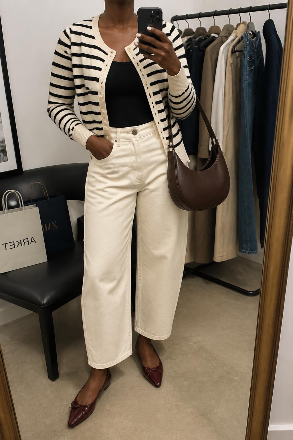 mirror selfie of a Black woman wearing a cream and black striped button-front cardigan buttoned halfway over a black fitted scoop-neck top, ecru high-rise barrel-leg jeans stopping above burgundy pointed-toe ballet flats, and a chocolate crescent shoulder bag, clearly showing the exact clothing items, colors, striped pattern, fabrics, shoes, and bag listed in the Outfit Pieces section, holding a phone in front of a mirror, cropped at NECK so the face does not show, relaxed casual selfie pose in a fashion studio mirror corner, soft indoor lighting, with a clothing rack holding a few garments behind her and shopping bags beside a chair, realistic knit, denim, leather-look, and structured bag textures, latest iPhone photo quality