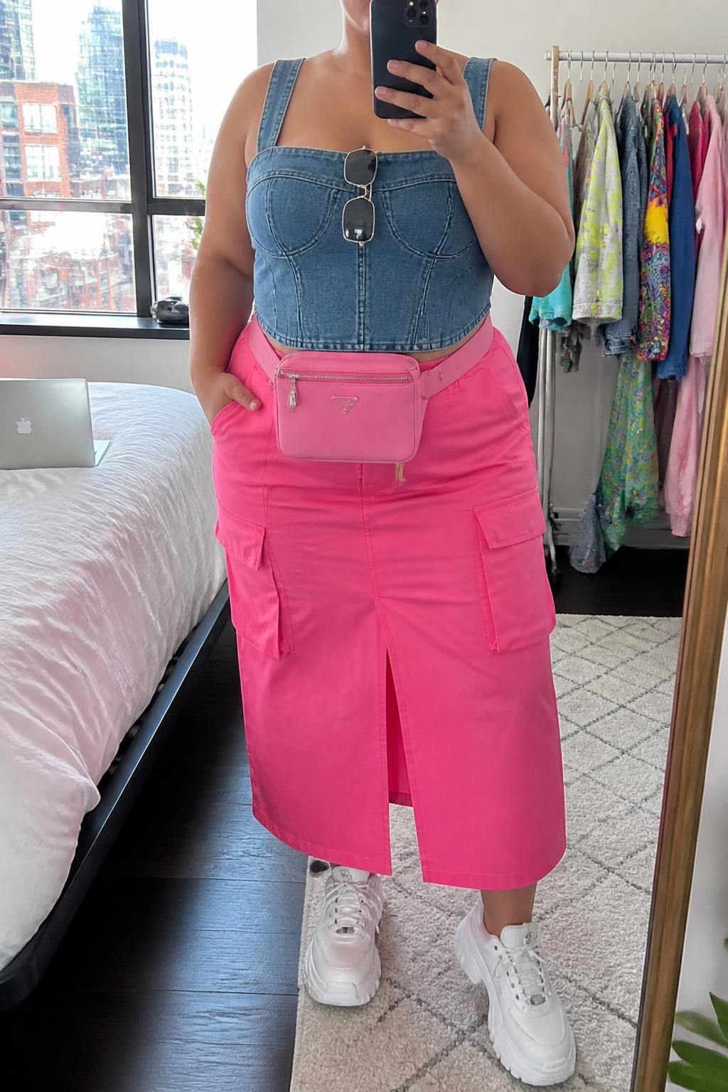 Mirror selfie of a plus size woman wearing a structured medium-wash denim corset top with wide straps, a bright pink cargo midi skirt with flat utility pockets and a front slit, white chunky sneakers, a small pink nylon belt bag, and silver rectangular sunglasses resting on the top edge of the corset, clearly showing the exact clothing items, colors, denim, pockets, slit, footwear, and accessories listed, holding a phone in front of a mirror, cropped at the neck so her face is hidden, relaxed confident selfie stance, in a stylish city apartment with natural window light, a laptop on the bed and a clothing rack with garments behind her, realistic bright festival styling, latest iPhone photo quality.