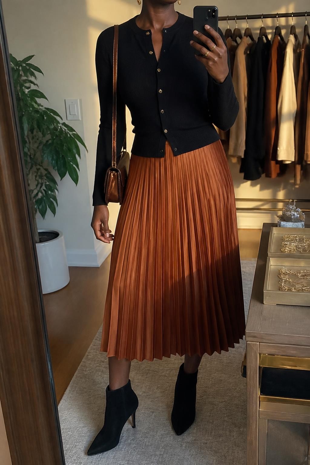 Mirror selfie of a Black woman wearing a fitted black cardigan with small gold buttons, a copper-orange pleated midi skirt, black suede ankle boots, a metallic bronze mini shoulder bag, and small gold drop earrings, clearly showing the exact clothing items, colors, fabrics, and accessories listed, holding a phone in front of a fashion studio mirror corner, cropped at the neck so the face does not show, relaxed casual selfie pose, evening golden-hour lighting, a clothing rack with garments behind and a jewelry tray on the vanity, polished natural setting, latest iPhone photo quality.