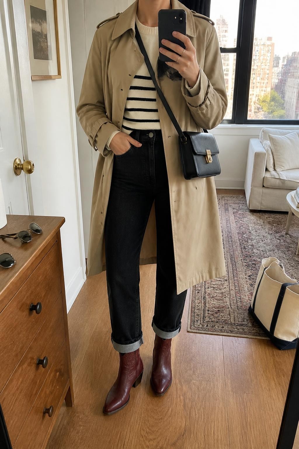 Image Prompt mirror selfie of a woman wearing an open camel water-resistant cotton trench coat over a cream and black striped fine-knit sweater, black high-rise straight-leg jeans rolled once above burgundy leather cowboy boots, and a black structured crossbody bag, clearly showing the exact clothing items, colors, striped pattern, fabrics, boots, and bag listed in the Outfit Pieces section, holding a phone in front of a mirror, cropped at NECK so the face does not show, relaxed travel-ready selfie pose in a stylish city apartment, natural window light, with a tote bag resting on the floor and sunglasses placed on a dresser, realistic cotton, fine knit, denim, and leather textures, latest iPhone photo quality