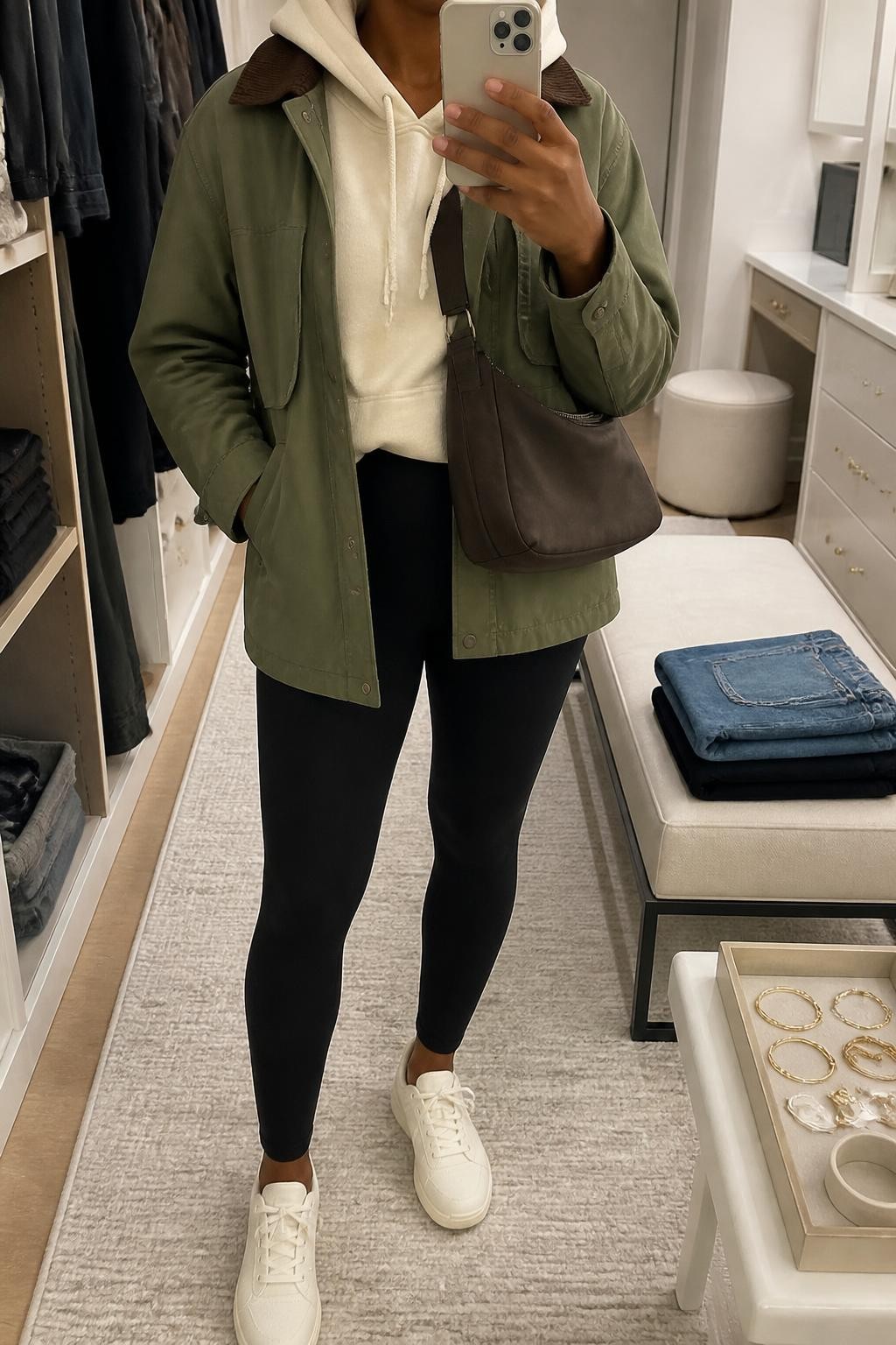 mirror selfie of a Black woman wearing an open olive cotton barn jacket with a brown corduroy collar over a cream brushed-cotton hoodie with the hood resting outside the jacket, black high-waisted ponte leggings, cream leather sneakers, and a chocolate nylon crossbody bag resting near the waist, clearly showing the exact clothing items, colors, fabrics, sneakers, and bag listed in the Outfit Pieces section, holding a phone in front of a mirror, cropped at NECK so the face does not show, relaxed casual selfie pose in a walk-in closet mirror, soft indoor lighting, with folded denim jeans on a bench and a jewelry tray on the vanity, realistic cotton, corduroy, fleece, ponte, leather, and nylon textures, latest iPhone photo quality