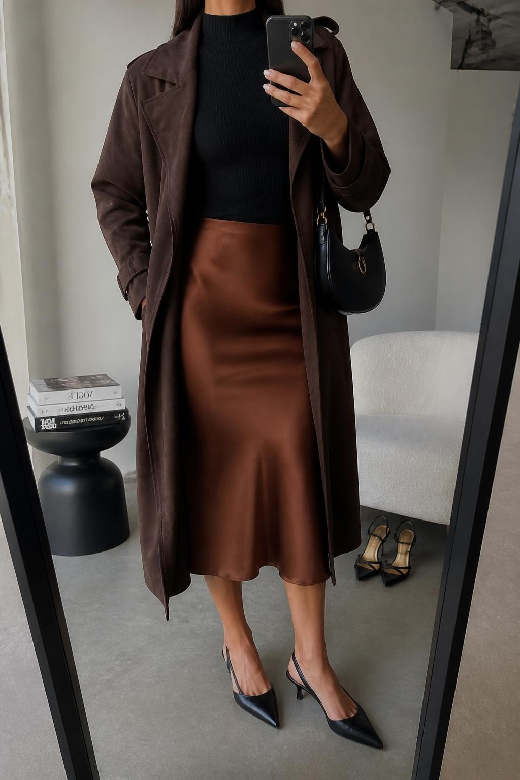 mirror selfie of a woman wearing an open chocolate brown faux-suede trench coat over a black fitted mock-neck knit top tucked into a copper satin midi slip skirt, black pointed-toe slingback heels, and a black crescent shoulder bag, clearly showing the exact clothing items, colors, fabrics, shoes, and bag listed in the Outfit Pieces section, holding a phone in front of a mirror, cropped at NECK so the face does not show, relaxed elegant selfie pose in a fashion studio mirror corner, soft indoor lighting, with a stack of fashion magazines on a side table and a pair of heels on the floor nearby, realistic faux-suede, knit, satin, and leather textures, latest iPhone photo quality