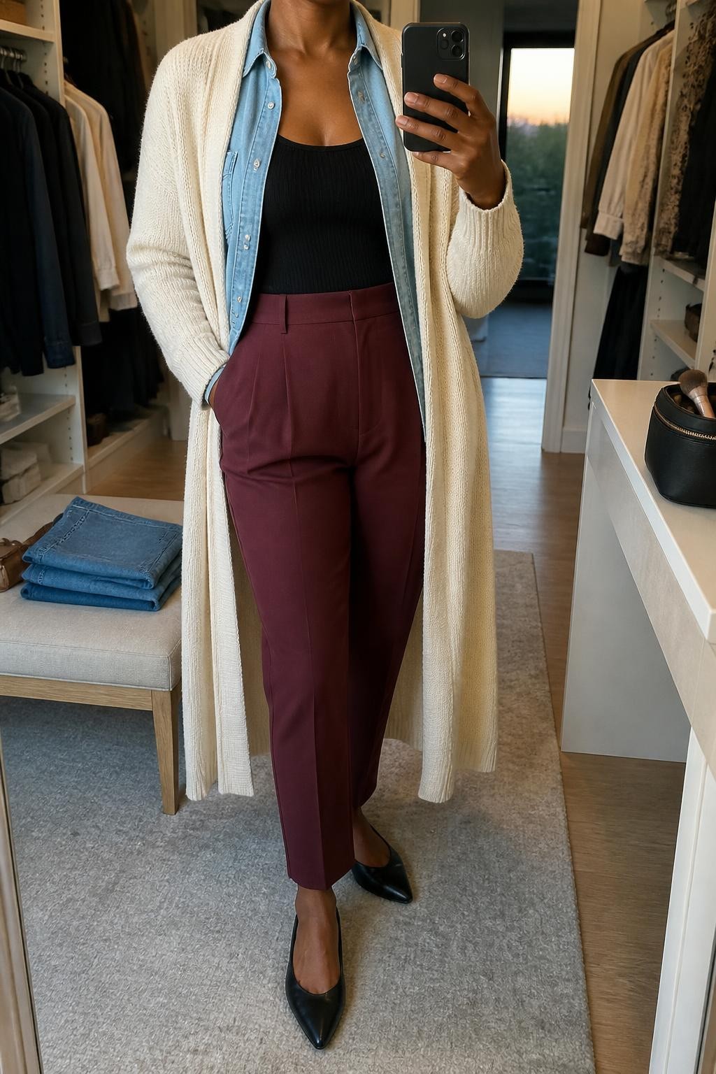 mirror selfie of a Black woman wearing an open cream longline duster cardigan over an open light-wash denim button-down shirt layered above a black fitted scoop-neck knit top, burgundy high-waisted tapered trousers, and black pointed-toe leather flats, clearly showing the exact clothing items, colors, fabrics, and shoes listed in the Outfit Pieces section, holding a phone in front of a mirror, cropped at NECK so the face does not show, relaxed professional selfie pose in a walk-in closet mirror, evening golden hour light, with folded denim jeans on a bench and a cosmetic bag on the vanity, realistic cardigan knit, denim, fitted knit, trouser, and leather textures, latest iPhone photo quality
