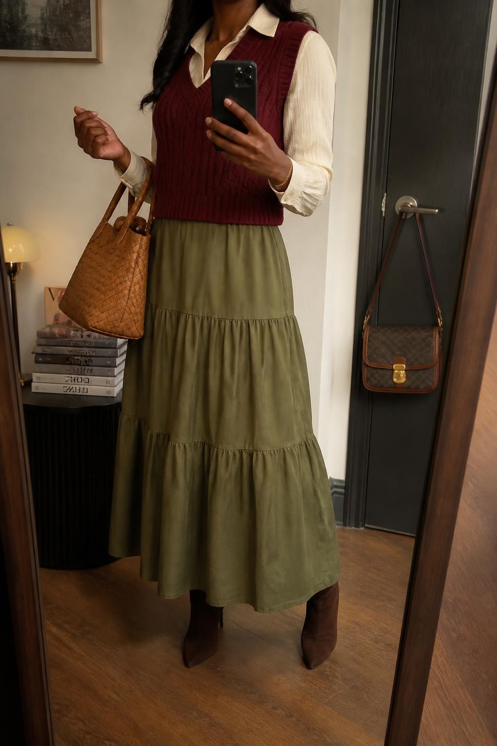 Mirror selfie of a Black woman wearing an olive-green tiered maxi skirt, a fitted cream long-sleeve blouse, a burgundy cable-knit vest, dark brown suede ankle boots, and a tan woven leather handbag, clearly showing the exact clothing items, colors, fabrics, and accessories listed, holding a phone in front of a stylish city apartment mirror, cropped at the neck so the face does not show, relaxed casual selfie pose, warm moody lighting, a stack of fashion magazines on a side table and a handbag hanging on a door handle, realistic and uncluttered, latest iPhone photo quality.