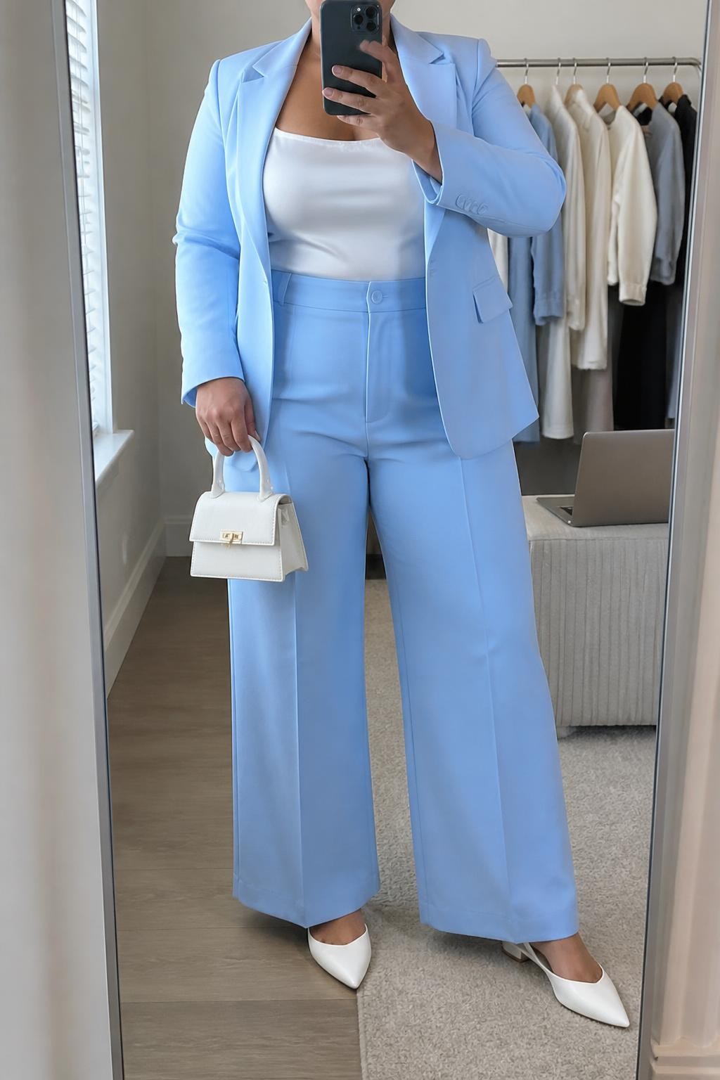 Mirror selfie of a plus size woman wearing a sky blue tailored blazer with matching high-waisted wide-leg trousers, a fitted white satin camisole with wide supportive straps, white pointed slingback flats, and a structured white mini handbag, clearly showing the exact suit, sky blue color, white satin, wide-leg shape, footwear, and accessories listed, holding a phone in front of a mirror, cropped at the neck so the face does not appear, relaxed confident selfie pose, in a minimal dressing room with natural window light, a clothing rack with garments behind her and a laptop resting on a bench, realistic modern baby shower outfit, latest iPhone photo quality.