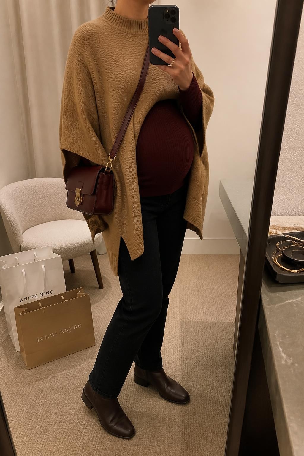 mirror selfie of a pregnant woman wearing a camel side-slit poncho sweater layered over a burgundy fitted ribbed maternity top shaped around her visible baby bump, black slim straight maternity jeans, dark brown flat leather ankle boots, and a burgundy structured crossbody bag, clearly showing the exact clothing items, colors, fabrics, boots, and bag listed in the Outfit Pieces section, holding a phone in front of a mirror, cropped at NECK so the face does not show, relaxed maternity selfie pose in a minimal dressing room, evening golden hour light, with shopping bags beside a chair and a jewelry tray on a nearby surface, realistic soft knit, ribbed fabric, denim, and leather textures, latest iPhone photo quality