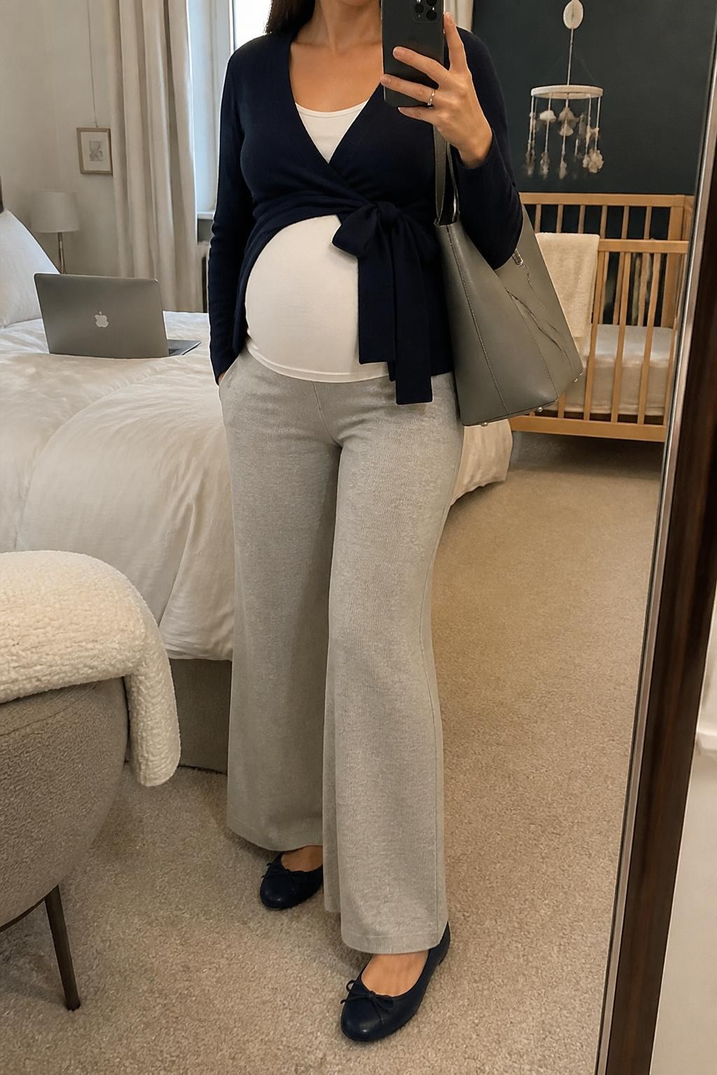 mirror selfie of a pregnant woman wearing a navy fine-knit maternity wrap cardigan tied slightly to one side above her visible baby bump over a white fitted maternity tank top, heather gray wide-leg knit maternity trousers, navy cushioned ballet flats, and a gray structured shoulder bag, clearly showing the exact clothing items, colors, fabrics, shoes, and bag listed in the Outfit Pieces section, holding a phone in front of a mirror, cropped at NECK so the face does not show, relaxed polished maternity selfie pose in a cozy bedroom mirror setup, soft indoor lighting, with a folded blanket on a chair and a laptop on the bed, realistic fine knit, soft trouser knit, and structured bag textures, latest iPhone photo quality