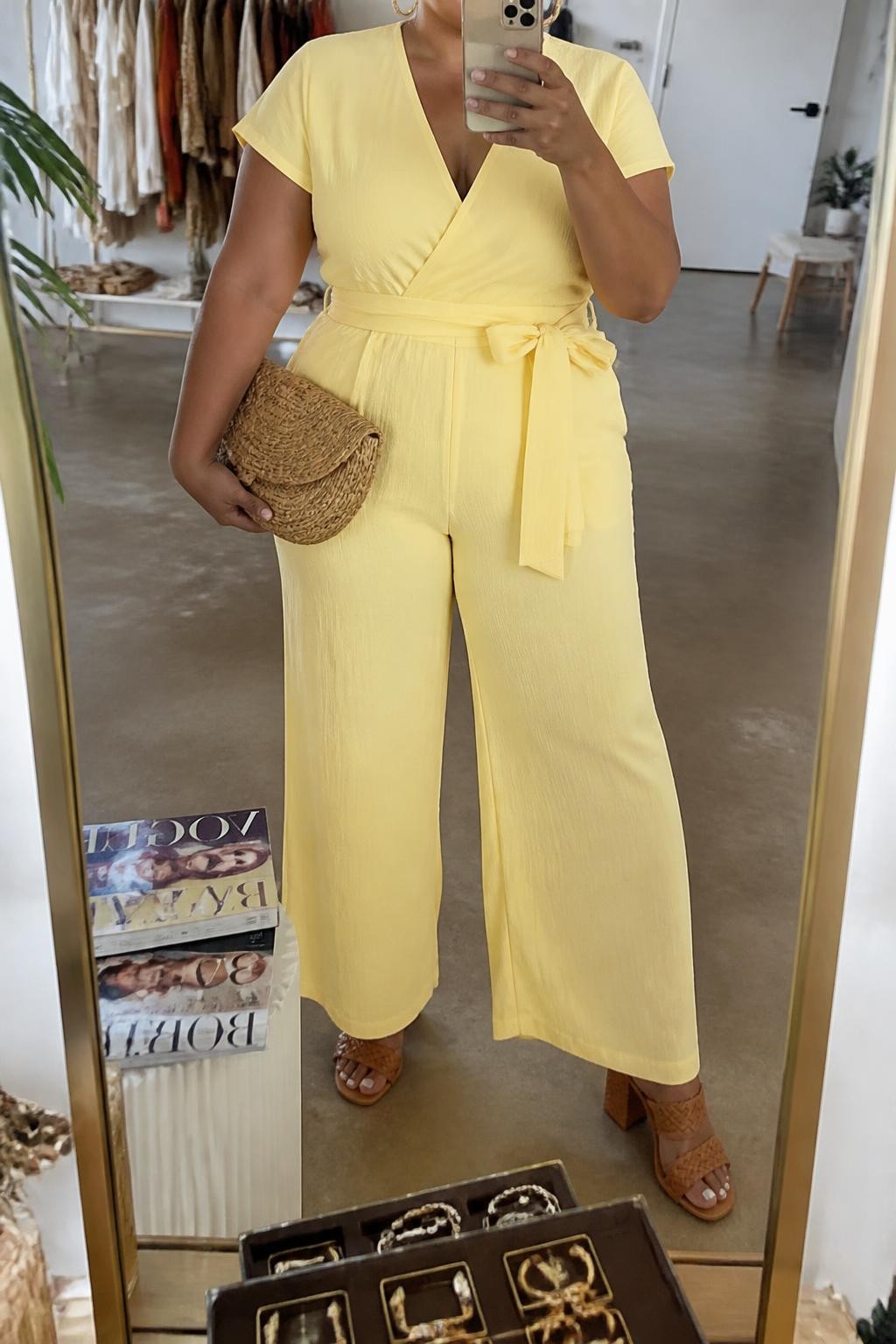 Mirror selfie of a plus size woman wearing a butter yellow wide-leg jumpsuit with short sleeves, a V-neckline, and a matching fabric belt, tan woven block-heel sandals, a natural raffia clutch, and hammered gold hoop earrings, clearly showing the exact jumpsuit, yellow color, waist belt, footwear, and accessories listed, holding a phone in front of a mirror, cropped at the neck so her face is hidden, relaxed cheerful selfie stance, in a fashion studio mirror corner with soft indoor lighting, a stack of fashion magazines on a side table and a jewelry tray on the vanity, realistic sunny brunch appearance, latest iPhone photo quality.