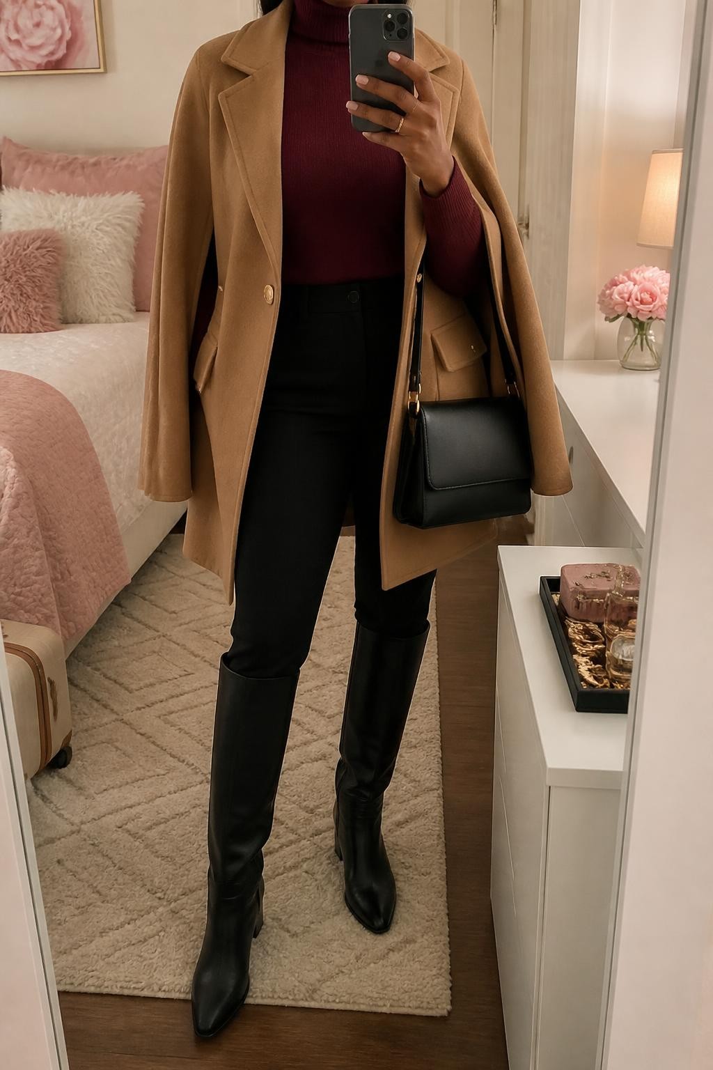 mirror selfie of a Black woman wearing a camel wool-blend cape coat partly open over a burgundy fitted fine-knit turtleneck, black slim straight-leg trousers tucked smoothly into black leather knee-high riding boots, and a black structured shoulder bag with a secure top zip, clearly showing the exact clothing items, colors, fabrics, boots, and bag listed in the Outfit Pieces section, holding a phone in front of a mirror, cropped at NECK so the face does not show, relaxed confident selfie pose in a feminine-themed bedroom, soft indoor lighting, with a jewelry tray on a dresser and a small suitcase beside the bed, realistic wool, fine knit, trouser fabric, and leather textures, latest iPhone photo quality