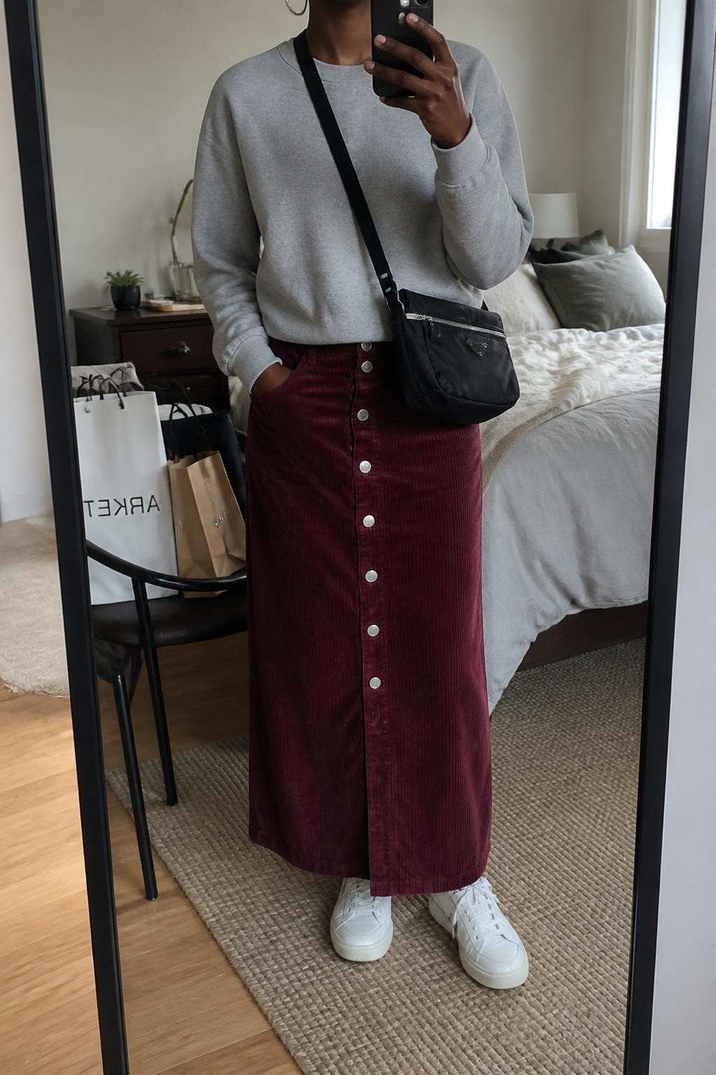 Mirror selfie of a Black woman wearing a burgundy corduroy maxi skirt with front buttons, soft grey crew-neck sweatshirt, white leather low-top sneakers, black nylon crossbody bag, and small silver hoop earrings, clearly showing the exact clothing items, colors, fabrics, and accessories listed, holding a phone in front of a cozy bedroom mirror setup, cropped at the neck so the face does not show, relaxed casual selfie pose, natural window light, soft pillows on an unmade bed and shopping bags beside a chair, casual believable environment, latest iPhone photo quality.