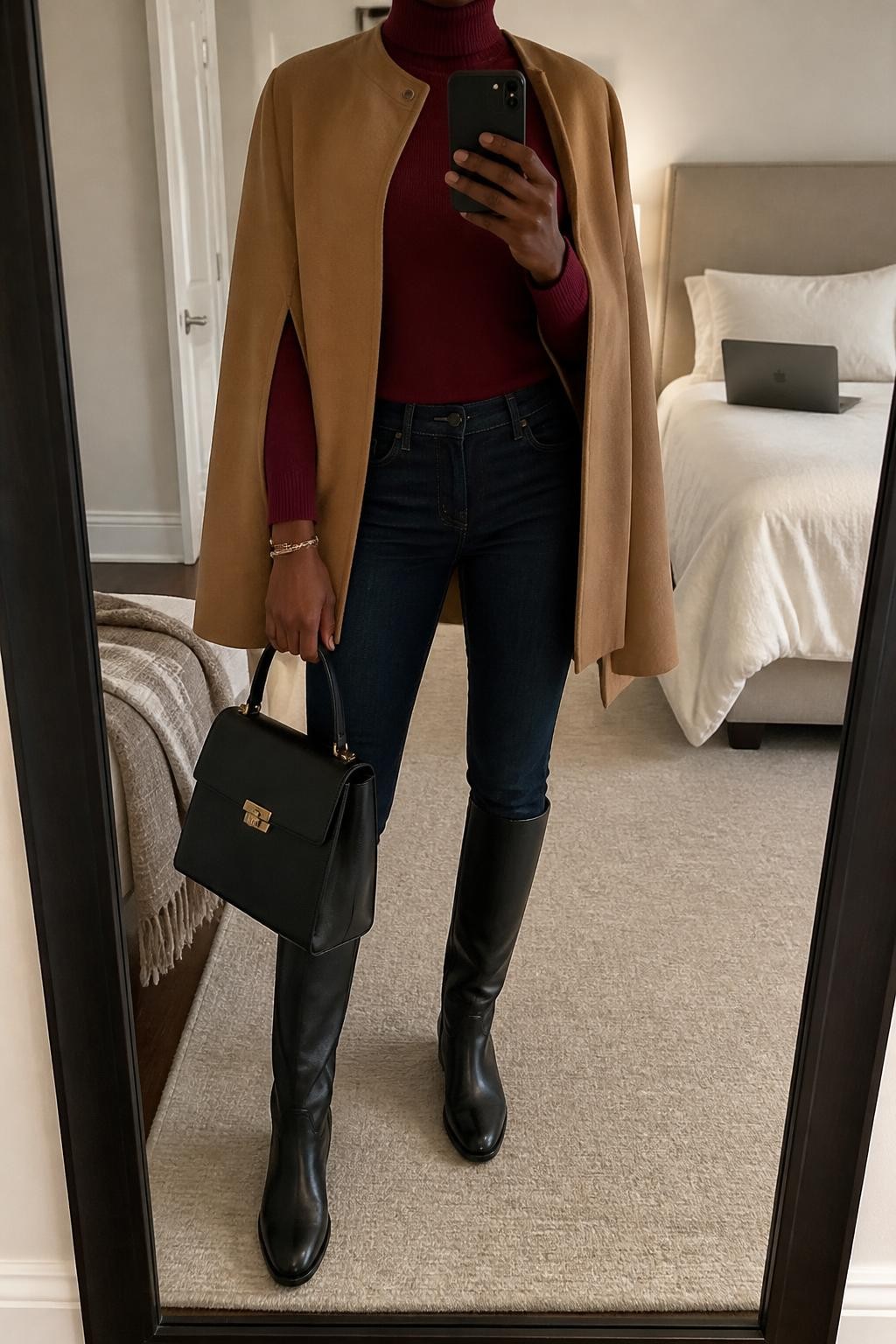 mirror selfie of a Black woman wearing a camel wool-blend cape coat left partly open over a burgundy fitted fine-knit turtleneck, dark indigo slim straight-leg jeans tucked smoothly into black leather knee-high riding boots, and a black structured top-handle bag, clearly showing the exact clothing items, colors, fabrics, boots, and bag listed in the Outfit Pieces section, holding a phone in front of a mirror, cropped at NECK so the face does not show, relaxed confident selfie pose in a cozy bedroom mirror setup, soft indoor lighting, with a folded blanket on a chair and a laptop on the bed, realistic wool, fine knit, denim, and leather textures, latest iPhone photo quality