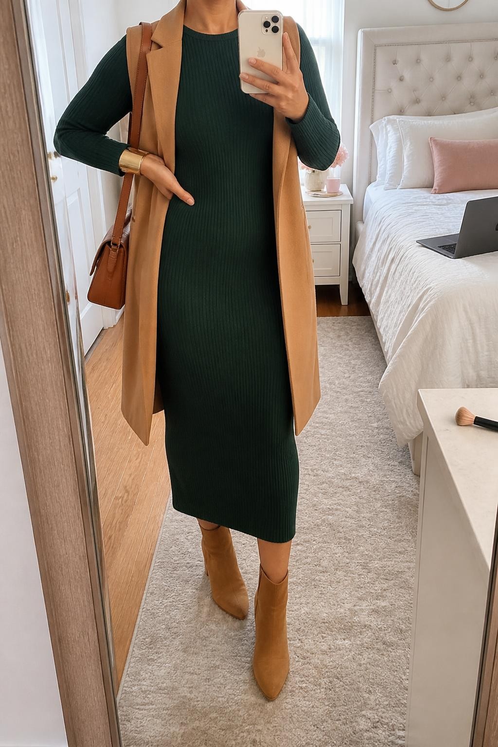 mirror selfie of a curvy woman wearing a forest green ribbed knit midi dress with a small space between the hem and tan suede block-heel ankle boots, an open camel longline tailored vest, a cognac leather shoulder bag, and a wide brushed gold bracelet, clearly showing the exact clothing items, colors, fabrics, boots, bag, and bracelet listed in the Outfit Pieces section, holding a phone in front of a mirror, cropped at NECK so the face does not show, relaxed polished selfie pose in a feminine-themed bedroom, natural window light, with a laptop on the bed and a makeup brush on a nearby counter, realistic ribbed knit, tailored fabric, suede, and leather textures, latest iPhone photo quality