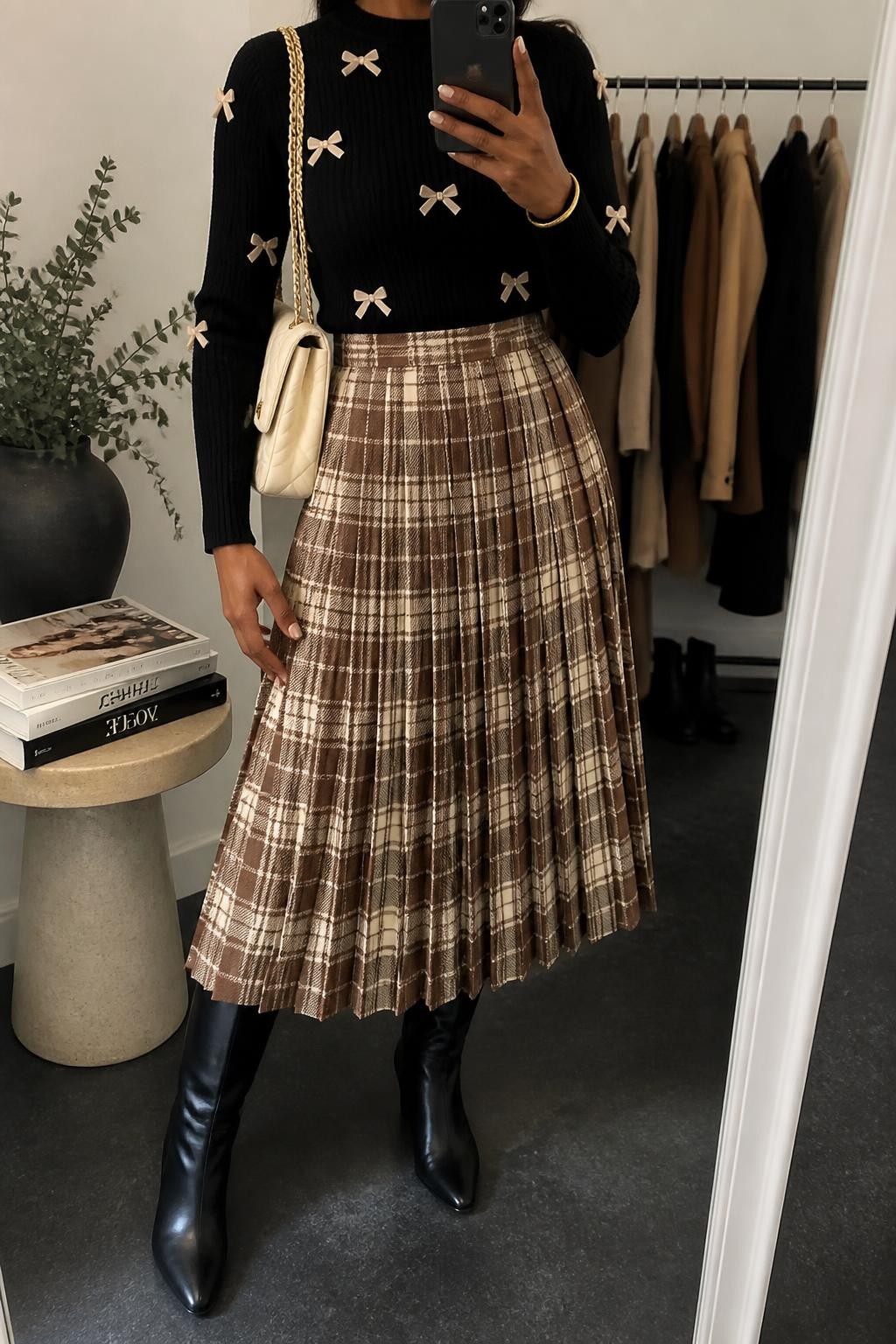 Image Prompt mirror selfie of a Black woman wearing a black fitted knit sweater with small cream velvet bows tucked fully into a brown and cream plaid pleated midi skirt, black leather knee-high boots, a cream quilted shoulder bag, and a slim gold cuff bracelet, clearly showing the exact clothing items, colors, plaid pattern, fabrics, bows, boots, bag, and bracelet listed in the Outfit Pieces section, holding a phone in front of a mirror, cropped at NECK so the face does not show, relaxed polished selfie pose in a fashion studio mirror corner, soft indoor lighting, with a stack of fashion magazines on a side table and a clothing rack with garments behind her, realistic knit, velvet, pleated fabric, leather, and quilted textures, latest iPhone photo quality