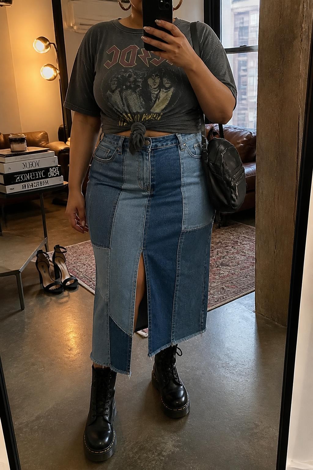 Mirror selfie of a plus size woman wearing a blue patchwork denim midi skirt with a front slit, an oversized faded charcoal graphic band T-shirt knotted gently at the waist, black lace-up combat boots, silver hoop earrings, and a small black nylon backpack, clearly showing the exact clothing items, colors, patchwork panels, graphic print, footwear, and accessories listed, holding a phone in front of a mirror, cropped at the neck so the face is hidden, natural relaxed selfie pose, in a stylish city apartment with warm moody lighting, a stack of fashion magazines on a side table and a pair of heels on the floor nearby, realistic vintage-inspired festival outfit, latest iPhone photo quality.