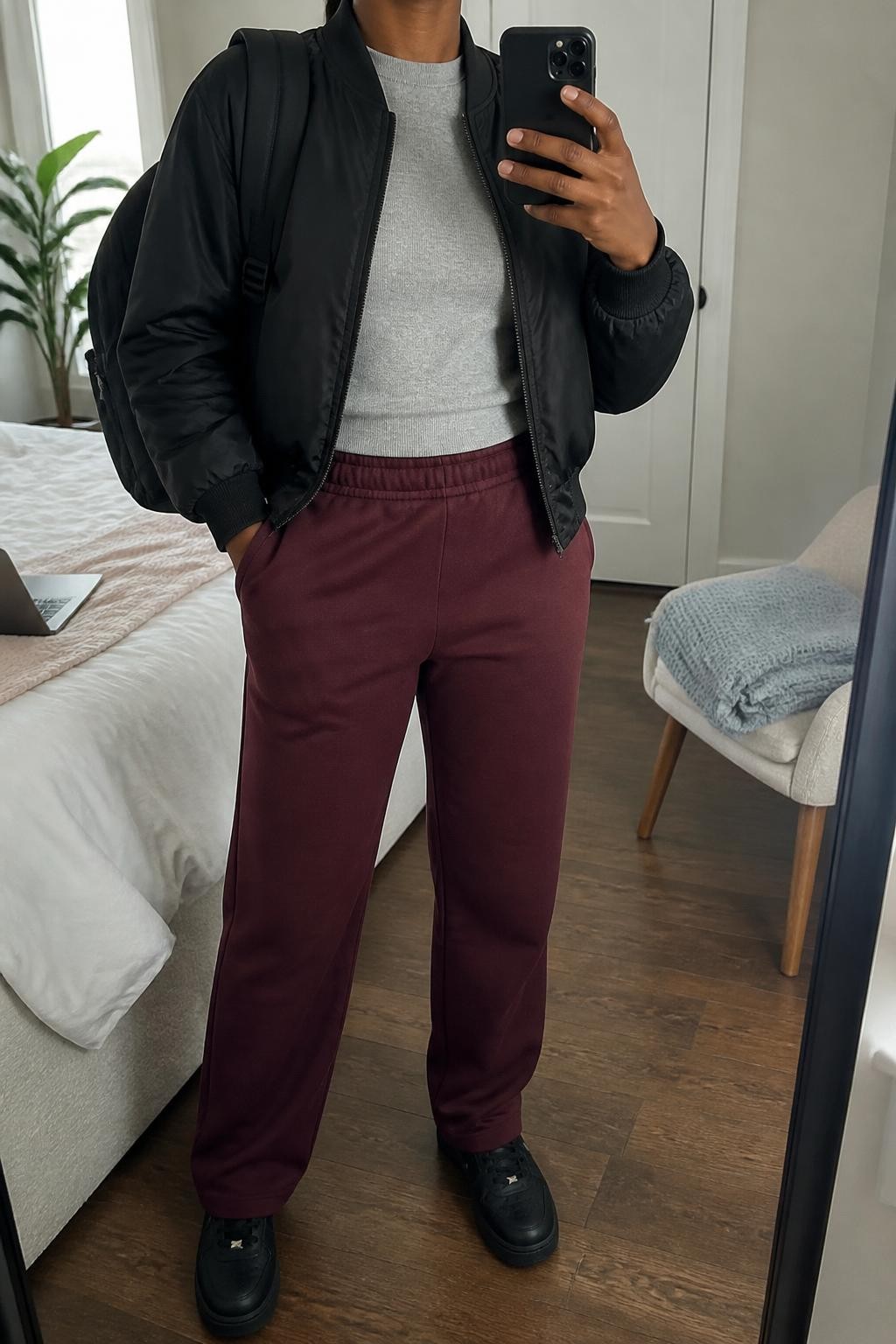 mirror selfie of a Black woman wearing an open black cropped nylon bomber jacket over a heather gray fitted crewneck sweatshirt, burgundy straight-leg joggers, black leather sneakers, and a black nylon backpack, clearly showing the exact clothing items, colors, fabrics, sneakers, and backpack listed in the Outfit Pieces section, holding a phone in front of a mirror, cropped at NECK so the face does not show, relaxed sporty selfie pose in a cozy bedroom mirror setup, natural window light, with a folded blanket on a chair and a laptop on the bed, realistic nylon, sweatshirt fabric, jogger fabric, and leather textures, latest iPhone photo quality