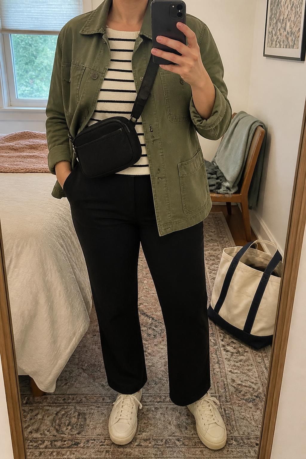 mirror selfie of a curvy woman wearing an olive cotton utility jacket with the sleeves pushed slightly above the wrists over a cream and black striped knit top, black straight-leg ponte trousers, cream leather sneakers, and a black nylon crossbody bag adjusted to sit near the waist, clearly showing the exact clothing items, colors, pattern, fabrics, sneakers, and bag listed in the Outfit Pieces section, holding a phone in front of a mirror, cropped at NECK so the face does not show, relaxed casual selfie pose in a cozy bedroom mirror setup, soft indoor lighting, with a folded blanket on a chair and a tote bag resting on the floor, realistic cotton, knit, ponte, leather, and nylon textures, latest iPhone photo quality