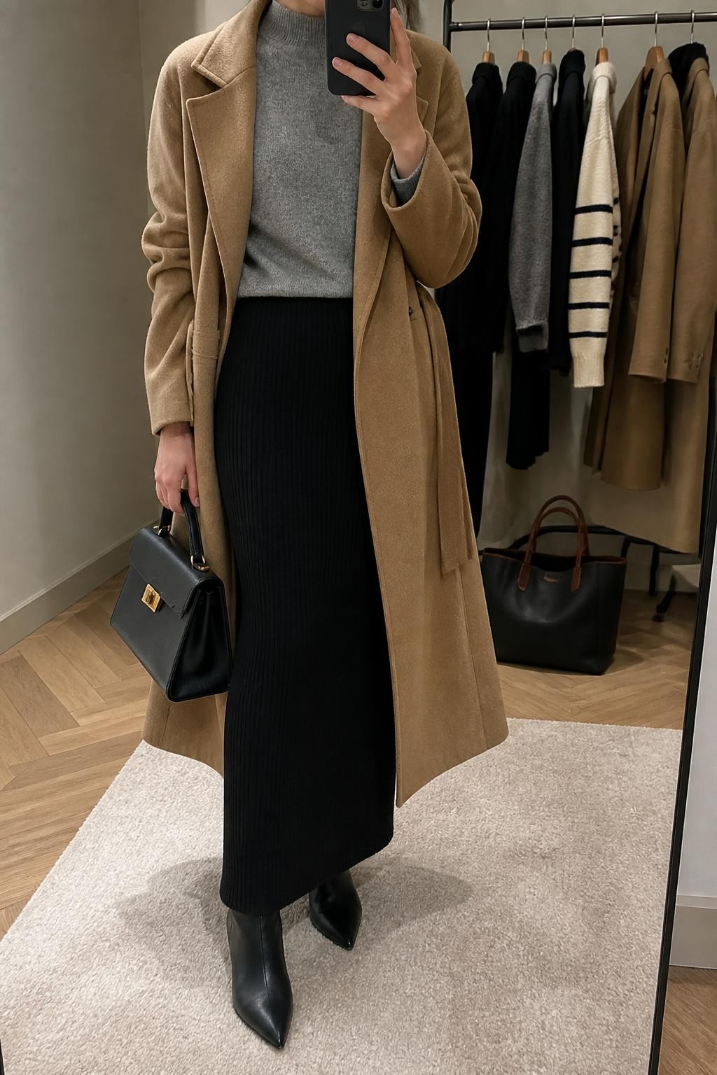 Mirror selfie of a woman wearing a fitted black ribbed-knit maxi skirt, camel wool wrap coat, soft grey mock-neck sweater, black pointed-toe ankle boots, and structured black leather handbag, clearly showing the exact clothing items, colors, fabrics, and accessories listed, holding a phone in front of a minimal dressing room mirror, cropped at the neck so the face does not show, relaxed casual selfie pose, soft indoor lighting, clothing rack with garments behind and a tote bag resting on the floor, polished but natural, latest iPhone photo quality.