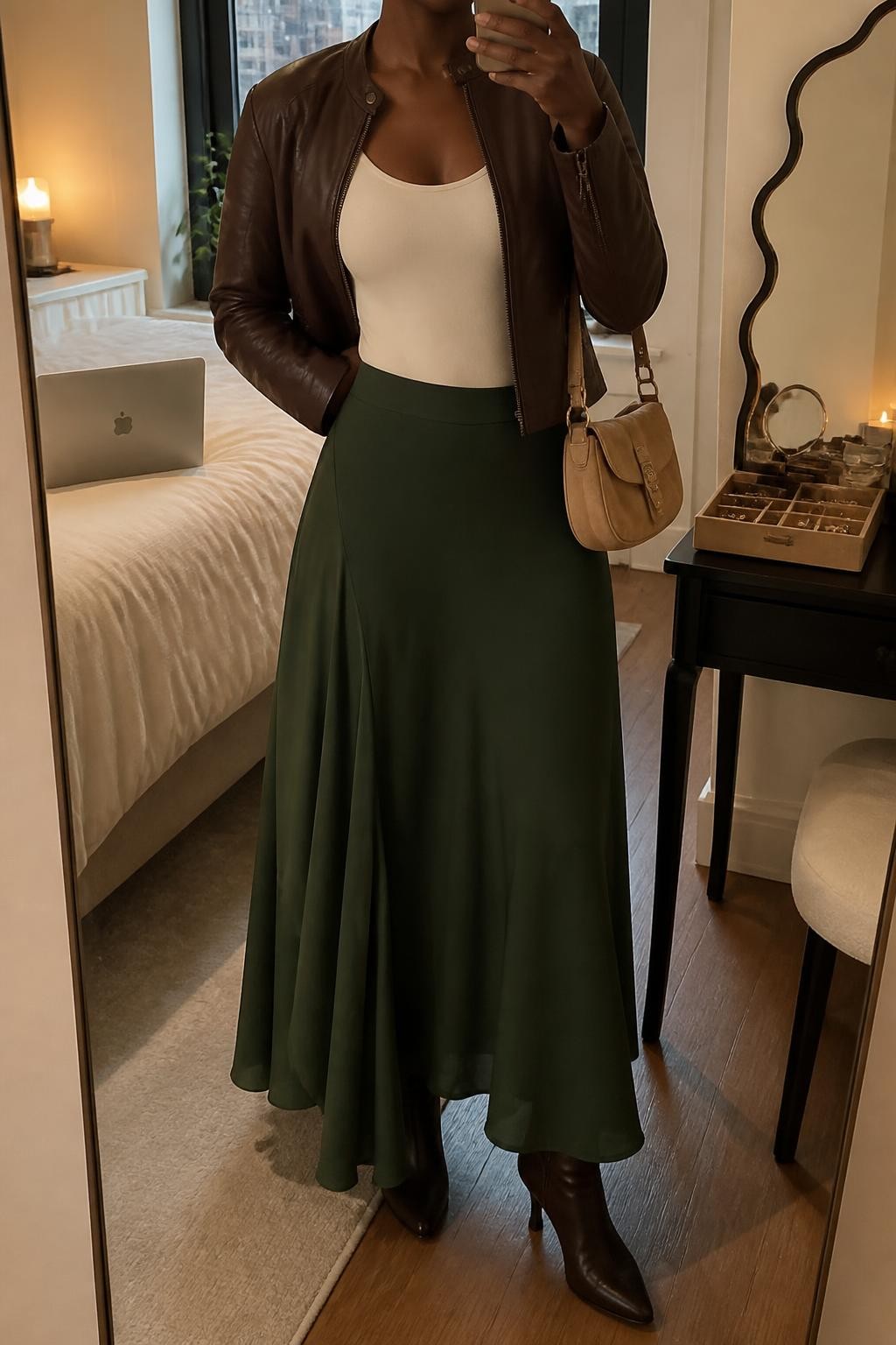 Mirror selfie of a Black woman wearing a flowing forest-green maxi skirt, fitted chocolate-brown leather jacket, cream scoop-neck bodysuit, dark brown heeled ankle boots, and small tan suede shoulder bag, clearly showing the exact clothing items, colors, fabrics, and accessories listed, holding a phone in front of a stylish city apartment mirror, cropped at the neck so the face does not show, relaxed casual selfie pose, warm moody lighting, laptop on the bed and a jewelry tray on the vanity, natural and casually styled, latest iPhone photo quality.