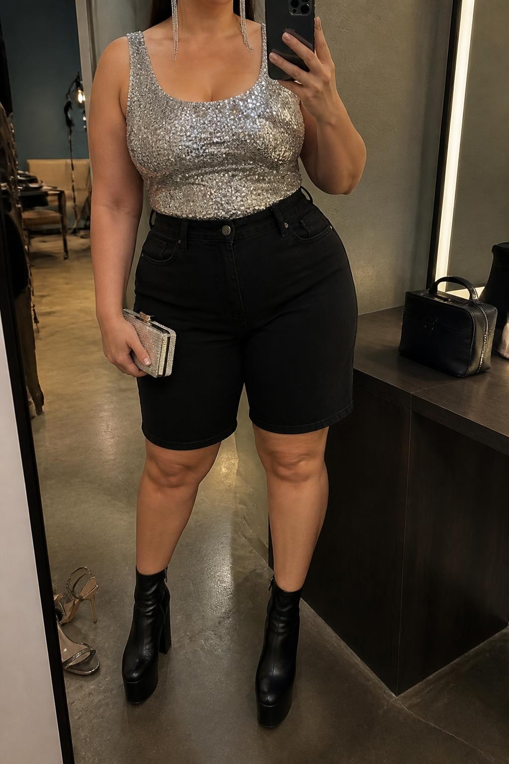 Mirror selfie of a plus size woman wearing black high-waisted denim jorts with a tailored knee-skimming cut, a fitted silver sequin sleeveless tank top with wide straps, black platform ankle boots, a small silver box clutch, and silver linear earrings, clearly showing the exact clothing items, black denim, silver sequins, footwear, and accessories listed, holding a phone in front of a mirror, cropped at the neck so the face is hidden, relaxed glamorous selfie stance, in a fashion studio mirror corner with warm moody lighting, a pair of heels on the floor nearby and a cosmetic bag on the counter, realistic summer party outfit, latest iPhone photo quality.