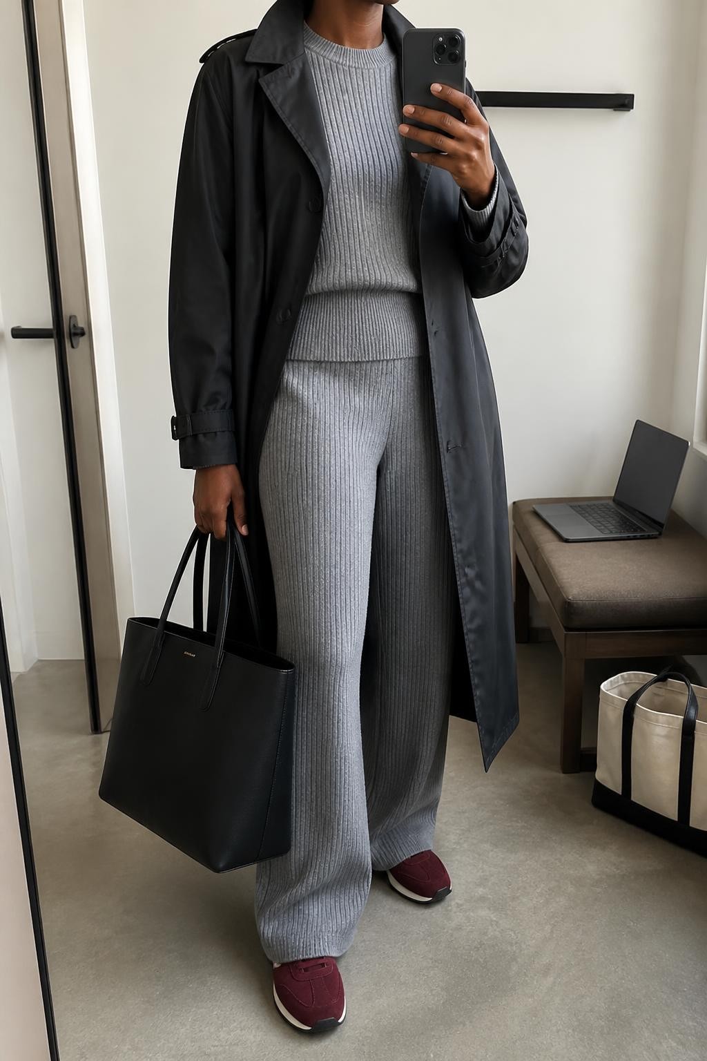 mirror selfie of a Black woman wearing an open black belted water-resistant trench coat over a matching heather gray ribbed crewneck sweater and heather gray ribbed wide-leg knit trousers, burgundy faux-suede sneakers, and a black structured travel tote, clearly showing the exact clothing items, colors, fabrics, sneakers, and bag listed in the Outfit Pieces section, holding a phone in front of a mirror, cropped at NECK so the face does not show, relaxed travel-ready selfie pose in a minimal dressing room, natural window light, with a laptop resting on a bench and a tote bag on the floor nearby, realistic water-resistant fabric, ribbed knit, faux-suede, and structured bag textures, natural casual mirror composition, latest iPhone photo quality