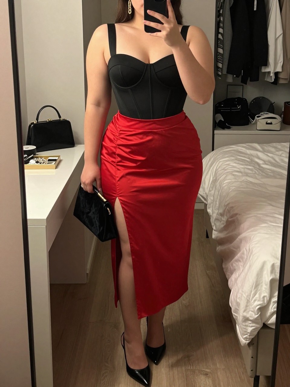 Mirror selfie of a plus size woman wearing a red high-waisted satin midi skirt with a front slit, a structured black corset-style bodysuit with wide supportive straps, black pointed slingback heels, a small black velvet clutch, and gold chain earrings, clearly showing the exact clothing items, red satin, black structured top, slit, footwear, and accessories listed, holding a phone in front of a mirror, cropped at the neck so the face is hidden, relaxed bold selfie pose, in a feminine-themed bedroom with warm moody lighting, a cosmetic bag on the counter and a jewelry tray on the vanity, realistic date-night outfit, latest iPhone photo quality.