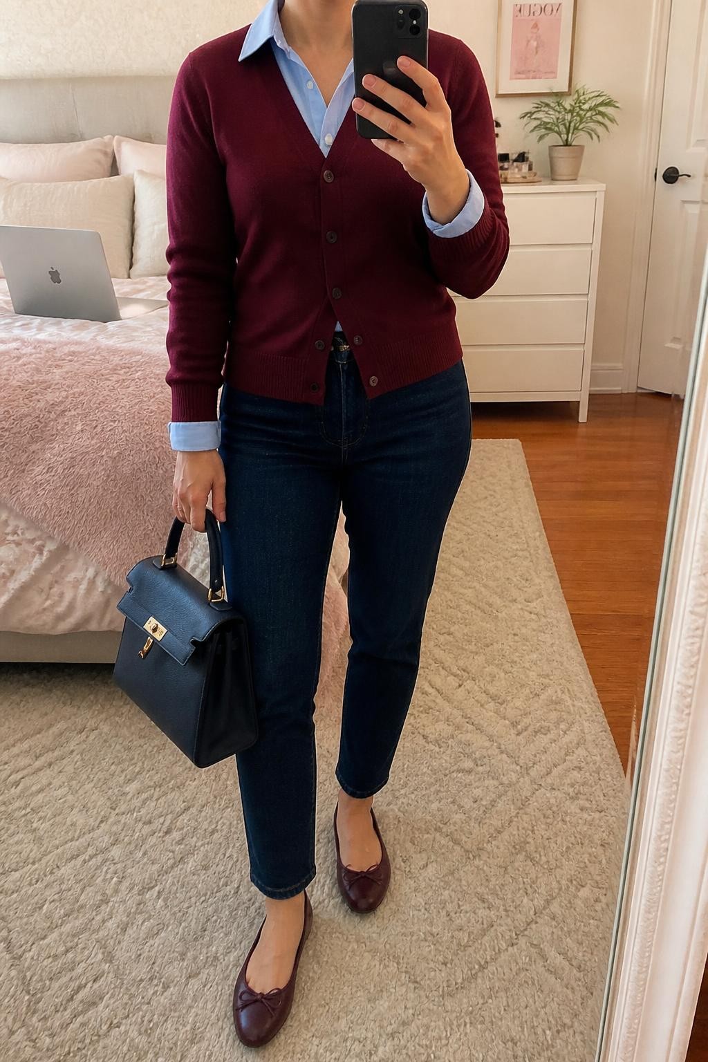 mirror selfie of a midsize woman wearing a burgundy V-neck knit cardigan over a powder blue cotton button-down shirt with the blue collar and cuffs visible, dark indigo cigarette jeans, burgundy leather ballet flats, and a navy structured top-handle bag, clearly showing the exact clothing items, colors, fabrics, shoes, and bag listed in the Outfit Pieces section, holding a phone in front of a mirror, cropped at NECK so the face does not show, relaxed casual selfie pose in a feminine-themed bedroom, soft indoor lighting, with a laptop resting on the bed and a small plant on a dresser, realistic cotton, knit, denim, and leather textures, natural everyday mirror photo, latest iPhone photo quality