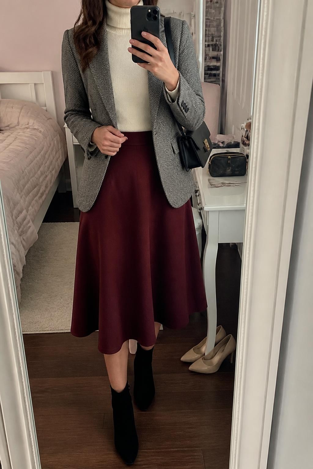 Mirror selfie of a woman wearing a deep burgundy A-line midi skirt, a fitted ivory turtleneck, a tailored grey herringbone blazer, black suede ankle boots, and a structured black shoulder bag, clearly showing the exact clothing items, colors, fabrics, and accessories listed, holding a phone in front of a feminine-themed bedroom mirror, cropped at the neck so the face does not show, relaxed confident selfie pose, soft indoor lighting, a pair of heels on the floor nearby and a makeup bag on the counter, natural and uncluttered, latest iPhone photo quality.