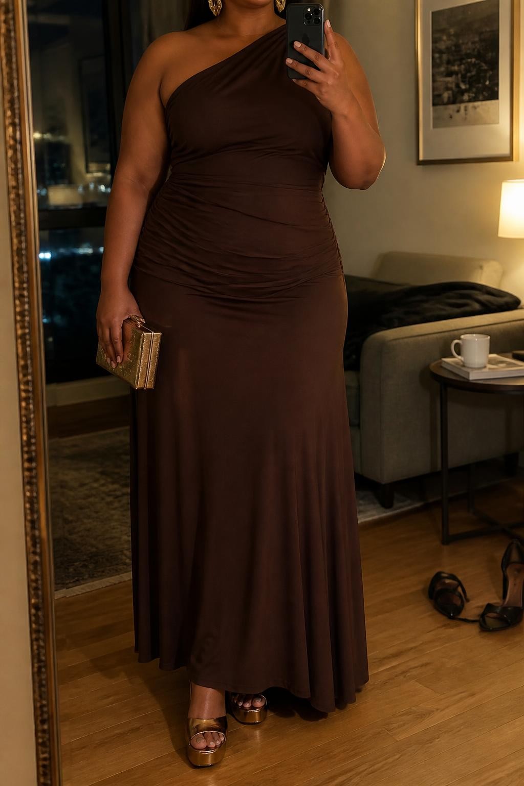 Mirror selfie of a plus size woman wearing a chocolate brown one-shoulder maxi dress in soft stretch jersey with gentle ruching across the waist and a flowing skirt, metallic bronze platform heels, a structured bronze box clutch, and chunky gold cuff earrings, clearly showing the exact dress shape, brown color, ruching, fabrics, footwear, and accessories listed, holding a phone in front of a mirror, cropped at the neck so the face does not show, relaxed confident selfie stance, in a stylish city apartment with warm moody lighting, a coffee mug on a side table and a pair of heels on the floor nearby, realistic upscale evening wedding appearance, latest iPhone photo quality.