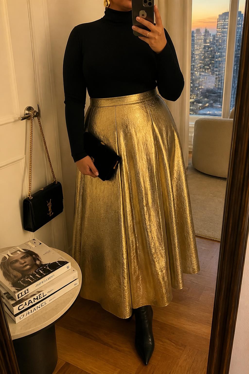 Mirror selfie of a plus size woman wearing a metallic gold high-waisted maxi skirt with a softly pleated shape, a fitted black long-sleeve turtleneck, black pointed ankle boots, a small black velvet clutch, and chunky gold cuff earrings, clearly showing the exact clothing items, gold metallic fabric, black knit, footwear, and accessories listed, holding a phone in front of a mirror, cropped at the neck so the face does not appear, relaxed elegant selfie pose, in a stylish city apartment with evening golden-hour lighting, a handbag hanging on a door handle and a stack of fashion magazines on a side table, realistic celebration outfit, latest iPhone photo quality.
