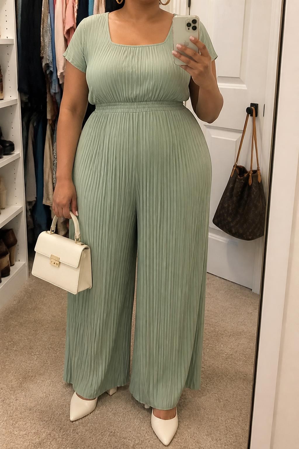 Mirror selfie of a plus size woman wearing a sage green pleated wide-leg jumpsuit with short sleeves, a square neckline, and a softly fitted elastic waist, cream pointed block heels, a structured cream top-handle bag, and delicate gold hoop earrings, clearly showing the exact jumpsuit, sage pleats, waist shape, cream footwear, and accessories listed, holding a phone in front of a mirror, cropped at the neck so her face does not appear, relaxed polished selfie stance, in a walk-in closet mirror with soft indoor lighting, an open closet with clothes visible and a handbag hanging on a door handle, realistic baby shower outfit, latest iPhone photo quality.