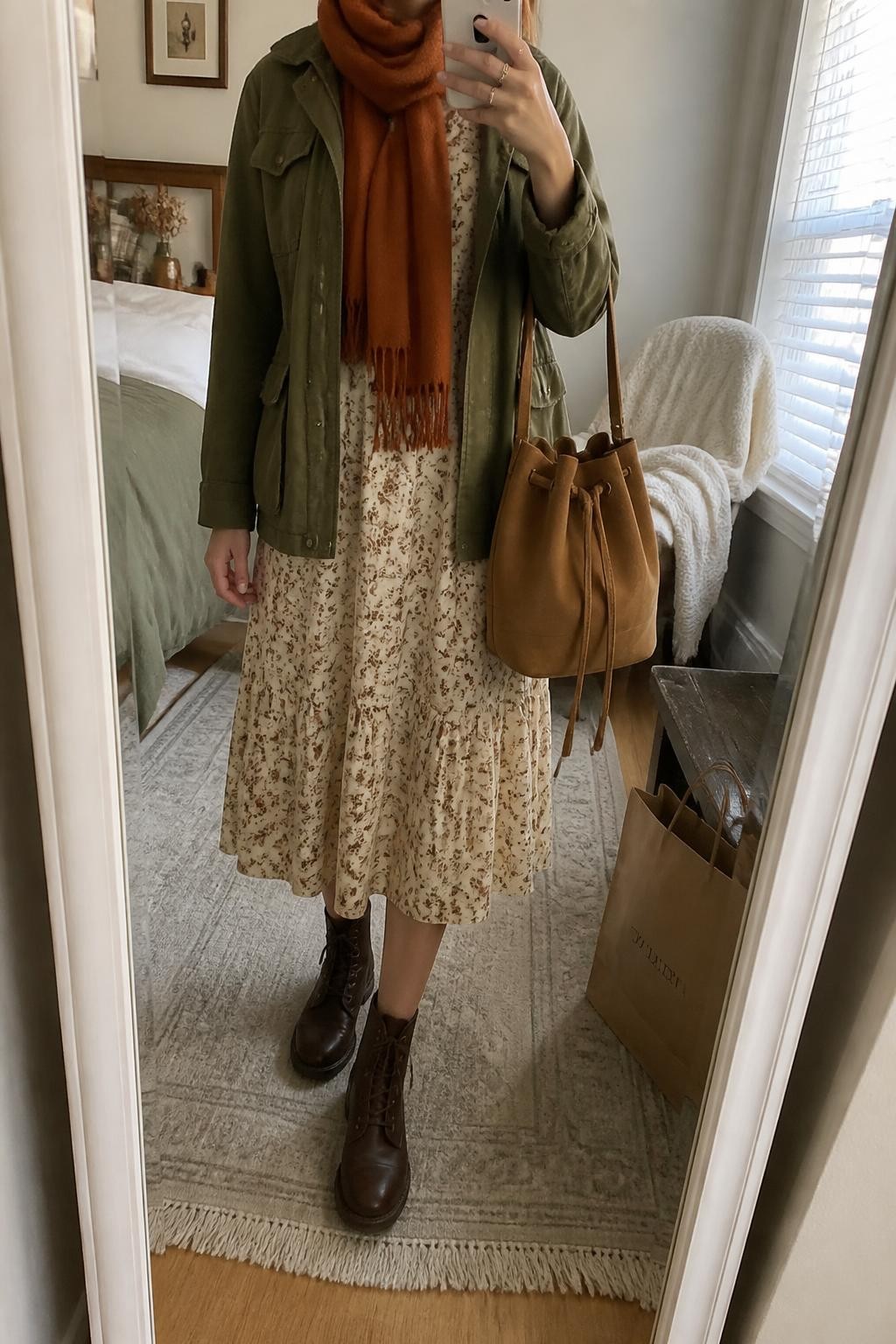 Mirror selfie of a woman wearing a cream cotton prairie midi dress with a small brown floral print, an olive-green utility jacket, chocolate-brown lace-up ankle boots, a tan suede bucket bag, and a rust-orange wool scarf, clearly showing the exact clothing items, colors, fabrics, and accessories listed, holding a phone in front of a cozy bedroom mirror setup, cropped at the neck so the face does not show, relaxed casual selfie pose, natural window light, a folded blanket on a chair and shopping bags beside a bench, believable everyday environment, latest iPhone photo quality.