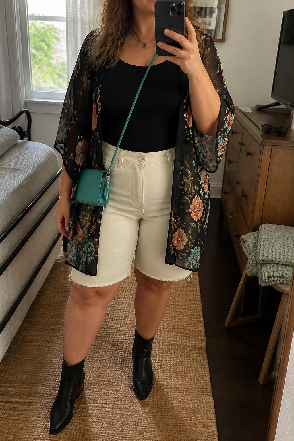 Mirror selfie of a plus size woman wearing a lightweight black chiffon kimono with coral, turquoise, and gold floral prints over a fitted black ribbed tank top, cream high-waisted denim jorts with a relaxed knee-skimming cut, black leather ankle boots, and a small turquoise crossbody bag, clearly showing the exact clothing layers, floral print, cream denim, black footwear, and accessories listed, holding a phone in front of a mirror, cropped at the neck so her face is hidden, relaxed festival selfie stance, in a cozy bedroom mirror setup with natural window light, sunglasses placed on a dresser and a folded blanket on a chair, realistic daytime festival outfit, latest iPhone photo quality.