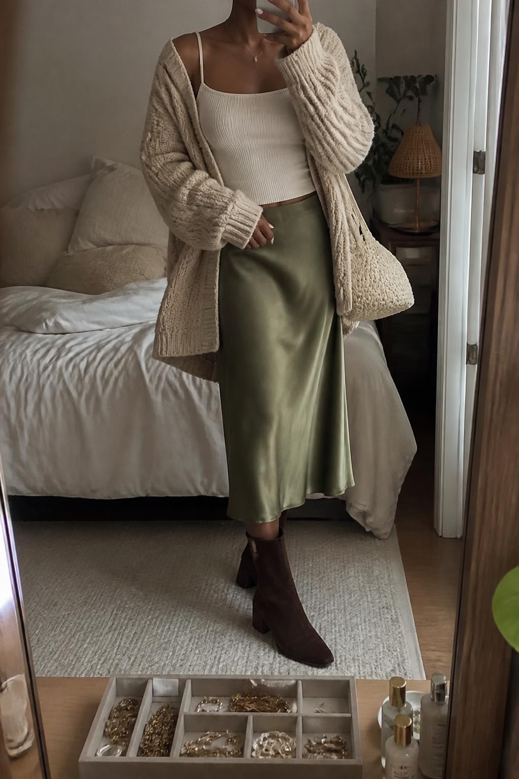 Mirror selfie of a Black woman wearing an oversized oatmeal chunky-knit cardigan over a fitted cream camisole, an olive-green satin midi skirt, dark brown suede ankle boots, and a cream woven shoulder bag, clearly showing the exact clothing items, colors, fabrics, and accessories listed, holding a phone in front of a cozy bedroom mirror setup, cropped at the neck so the face does not show, relaxed casual selfie pose, warm moody lighting, soft pillows on an unmade bed and a jewelry tray on the vanity, realistic lived-in setting, latest iPhone photo quality.