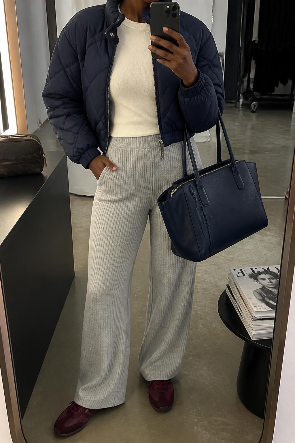 mirror selfie of a Black woman wearing an open navy cropped quilted puffer jacket over an ivory fitted crewneck sweater, heather gray ribbed wide-leg knit trousers, burgundy leather sneakers, and a navy structured travel tote, clearly showing the exact clothing items, colors, fabrics, sneakers, and bag listed in the Outfit Pieces section, holding a phone in front of a mirror, cropped at NECK so the face does not show, relaxed travel-ready selfie pose in a fashion studio mirror corner, soft indoor lighting, with a stack of fashion magazines on a side table and a cosmetic bag on the counter, realistic quilting, knit, leather, and structured bag textures, latest iPhone photo quality