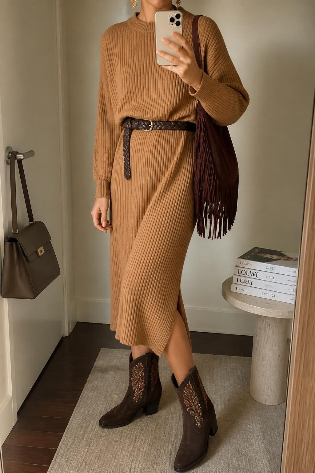 Mirror selfie of a woman wearing a loose camel ribbed-knit midi sweater dress, a dark brown braided leather belt, chocolate-brown suede ankle boots with rust and cream embroidery, a burgundy fringed shoulder bag, and antique-gold drop earrings, clearly showing the exact clothing items, colors, fabrics, and accessories listed, holding a phone in front of a minimal dressing room mirror, cropped at the neck so the face does not show, relaxed casual selfie pose, natural window light, a handbag hanging on a door handle and a stack of fashion magazines on a side table, warm natural environment, latest iPhone photo quality.