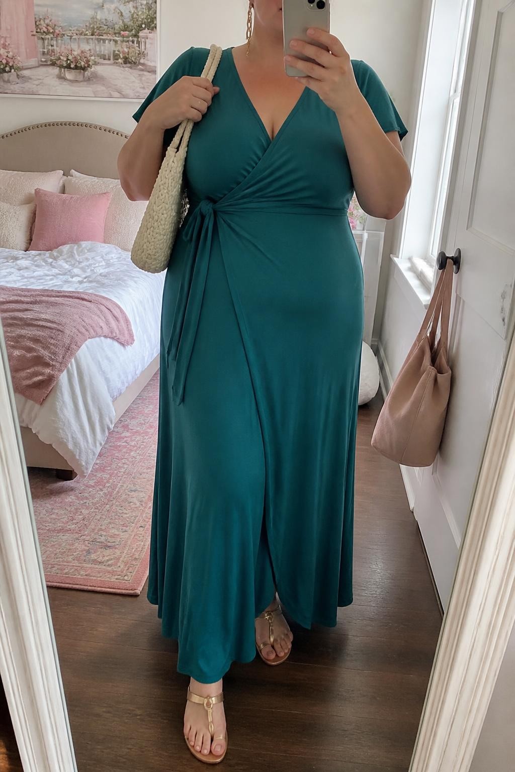 Mirror selfie of a plus size woman wearing a teal green jersey wrap maxi dress with short flutter sleeves, a modest V-neckline, and a softly tied waist, metallic gold flat toe-ring sandals, a cream woven shoulder bag, and delicate gold chain earrings, clearly showing the exact dress, teal jersey, wrap shape, footwear, and accessories listed, holding a phone in front of a mirror, cropped at the neck so the face is hidden, relaxed graceful selfie pose, in a feminine-themed bedroom with natural window light, soft pillows on an unmade bed and a handbag hanging on a door handle, realistic resort brunch styling, latest iPhone photo quality.