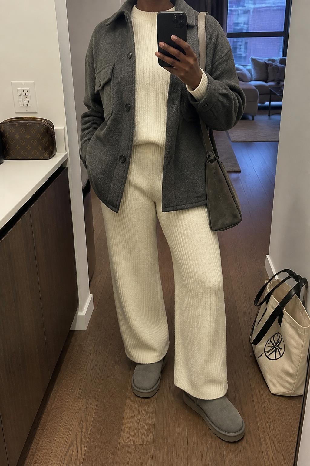 mirror selfie of a Black woman wearing an open charcoal brushed wool-blend shacket over a cream ribbed knit crewneck sweater, matching cream ribbed knit wide-leg trousers, heather gray cuffed Ugg boots, and a gray suede shoulder bag, clearly showing the exact clothing items, colors, fabrics, boots, and bag listed in the Outfit Pieces section, holding a phone in front of a mirror, cropped at NECK so the face does not show, relaxed casual selfie pose in a stylish city apartment, soft indoor lighting, with a tote bag resting on the floor and a cosmetic bag on a nearby counter, realistic brushed wool, ribbed knit, and suede textures, slightly imperfect mirror composition, latest iPhone photo quality