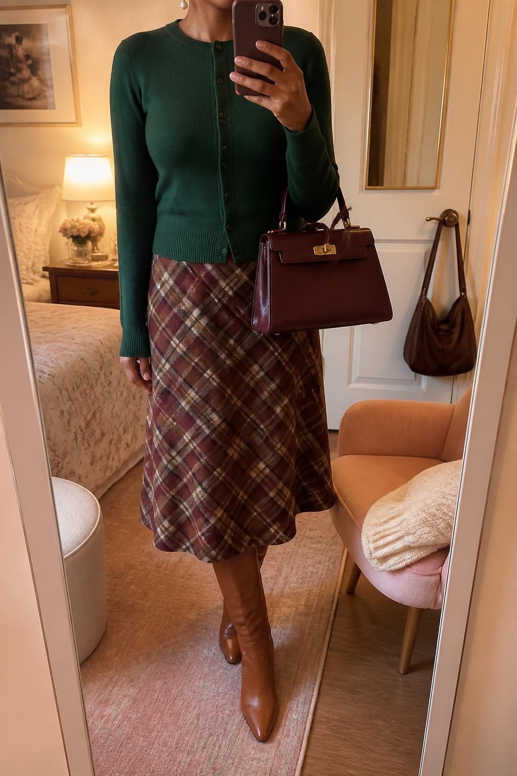 Mirror selfie of a Black woman wearing a fitted forest-green crew-neck cardigan, a burgundy, camel, and cream plaid wool midi skirt, cognac leather knee-high boots, a structured burgundy handbag, and small pearl drop earrings, clearly showing the exact clothing items, colors, fabrics, and accessories listed, holding a phone in front of a feminine-themed bedroom mirror, cropped at the neck so the face does not show, relaxed casual selfie pose, evening golden-hour lighting, a folded blanket on a chair and a handbag hanging on a door handle, warm realistic setting, latest iPhone photo quality.