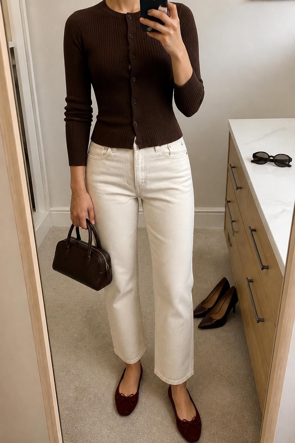 Image Prompt mirror selfie of a woman wearing a fully buttoned chocolate ribbed corset-style cardigan, cream high-rise straight-leg jeans stopping just above burgundy suede ballet flats with small bows, a chocolate structured mini handbag, and a thin gold bracelet, clearly showing the exact clothing items, colors, fabrics, buttons, bows, shoes, bag, and bracelet listed in the Outfit Pieces section, holding a phone in front of a mirror, cropped at NECK so the face does not show, relaxed casual selfie pose in a minimal dressing room, soft indoor lighting, with sunglasses placed on a dresser and a pair of heels on the floor nearby, realistic ribbed knit, denim, suede, leather, and metal textures, latest iPhone photo quality
