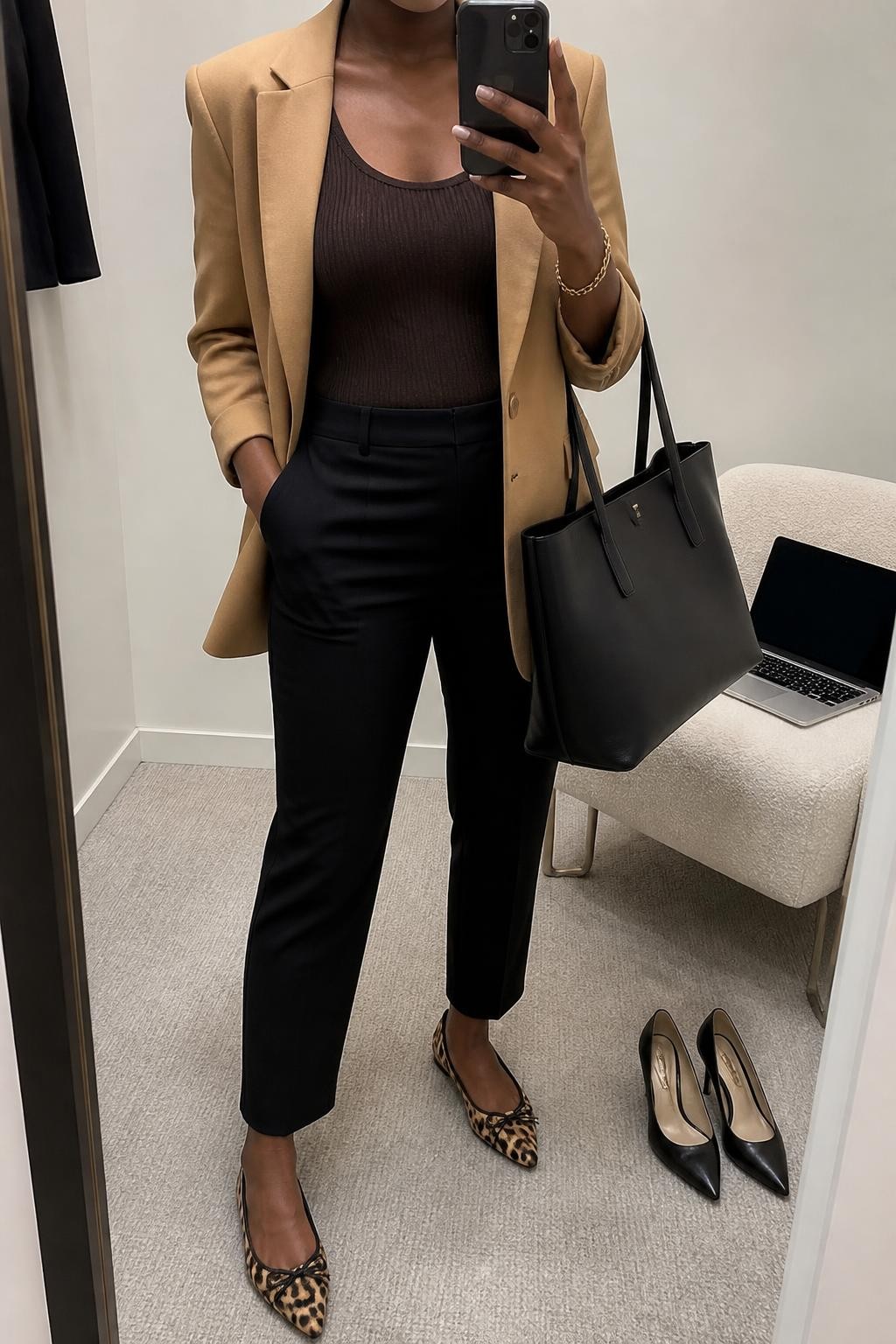 mirror selfie of a Black woman wearing an open camel relaxed-fit single-breasted blazer over a chocolate fitted scoop-neck knit top, black ankle-length tailored trousers, leopard-print pointed-toe ballet flats, and a black structured work tote, clearly showing the exact clothing items, colors, pattern, fabrics, shoes, and bag listed in the Outfit Pieces section, holding a phone in front of a mirror, cropped at NECK so the face does not show, relaxed professional selfie pose in a minimal dressing room, soft indoor lighting, with a laptop resting on a chair and a pair of heels on the floor nearby, realistic tailoring, knit, printed fabric, and leather-look textures, latest iPhone photo quality