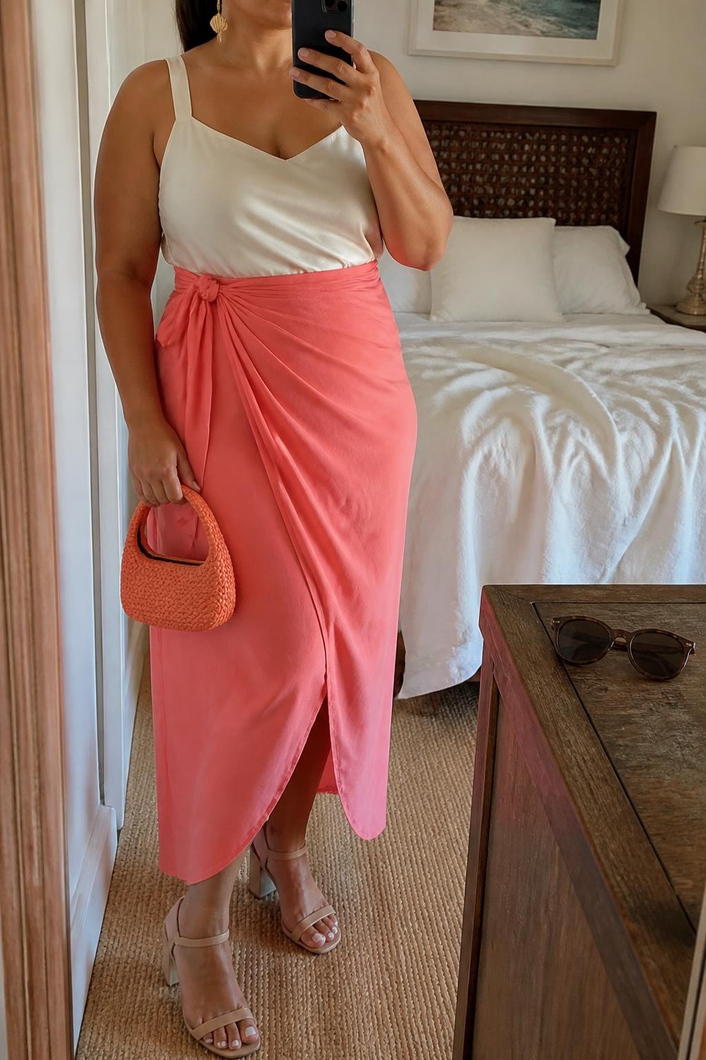 Mirror selfie of a plus size woman wearing a coral pink high-waisted wrap maxi skirt with a softly draped front and adjustable side tie, an ivory satin camisole with wide supportive straps, nude block-heel sandals, a coral woven mini bag, and delicate gold shell earrings, clearly showing the exact clothing items, coral wrap shape, ivory satin, footwear, and accessories listed, holding a phone in front of a mirror, cropped at the neck so her face is hidden, relaxed graceful selfie pose, in a cozy bedroom mirror setup with soft indoor lighting, soft pillows on an unmade bed and sunglasses placed on a dresser, realistic resort dinner styling, latest iPhone photo quality.