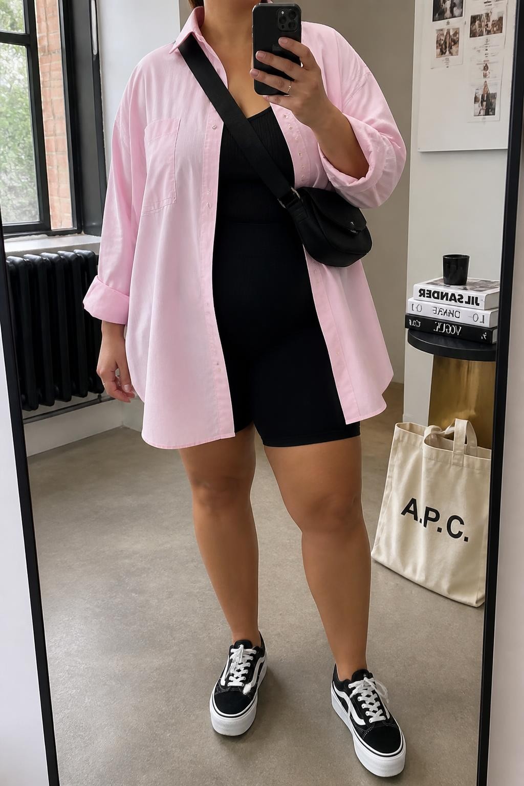 Mirror selfie of a plus size woman wearing an oversized soft pink cotton Oxford shirt with a long curved hem, black high-waisted biker shorts, a fitted black ribbed tank top beneath the shirt, black and white platform sneakers, and a compact black nylon crossbody bag, clearly showing the exact clothing layers, pink cotton, black shorts, footwear, and accessories listed, holding a phone in front of a mirror, cropped at the neck so her face is hidden, relaxed sporty selfie stance, in a fashion studio mirror corner with natural window light, a tote bag resting on the floor and a stack of fashion magazines on a side table, realistic weekend outfit, latest iPhone photo quality.