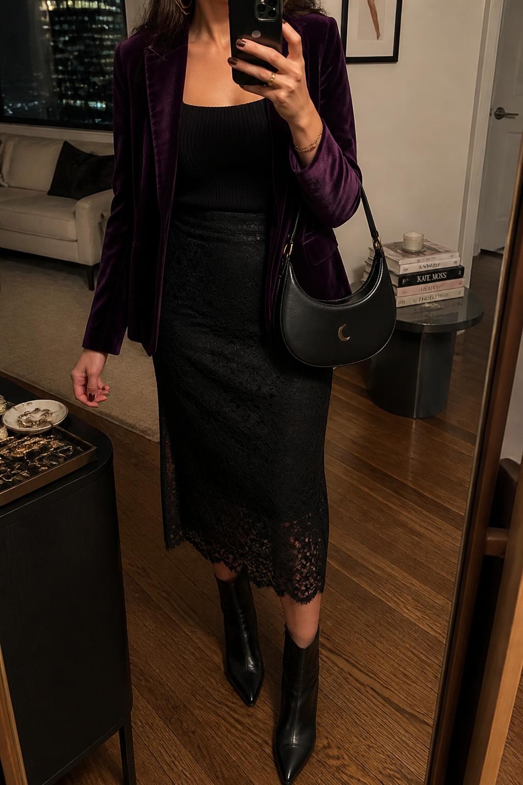 mirror selfie of a woman wearing an open deep plum fitted velvet blazer over a black ribbed square-neck knit top tucked into a black lace-overlay midi skirt, black pointed-toe leather ankle boots, and a black crescent bag with a silver moon clasp, clearly showing the exact clothing items, colors, fabrics, boots, bag, and clasp listed in the Outfit Pieces section, holding a phone in front of a full-length mirror, cropped at NECK so the face does not show, relaxed confident selfie pose in a stylish city apartment, warm moody lighting, with a stack of fashion magazines on a side table and a jewelry tray on a dresser, realistic velvet, ribbed knit, lace, and leather textures, natural mirror reflection, latest iPhone photo quality