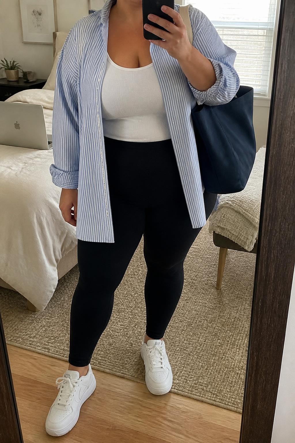 Mirror selfie of a plus size woman wearing an oversized blue and white striped cotton button-down shirt worn open over a fitted white ribbed scoop-neck tank top, black high-waisted stretch leggings, white cushioned leather sneakers, and a large navy canvas tote bag, clearly showing the exact clothing layers, stripe pattern, black leggings, white footwear, and accessories listed, holding a phone in front of a mirror, cropped at the neck so her face does not appear, relaxed travel-ready selfie stance, in a cozy bedroom mirror setup with natural window light, a laptop resting on the bed and a folded blanket on a chair, realistic casual outfit, latest iPhone photo quality.