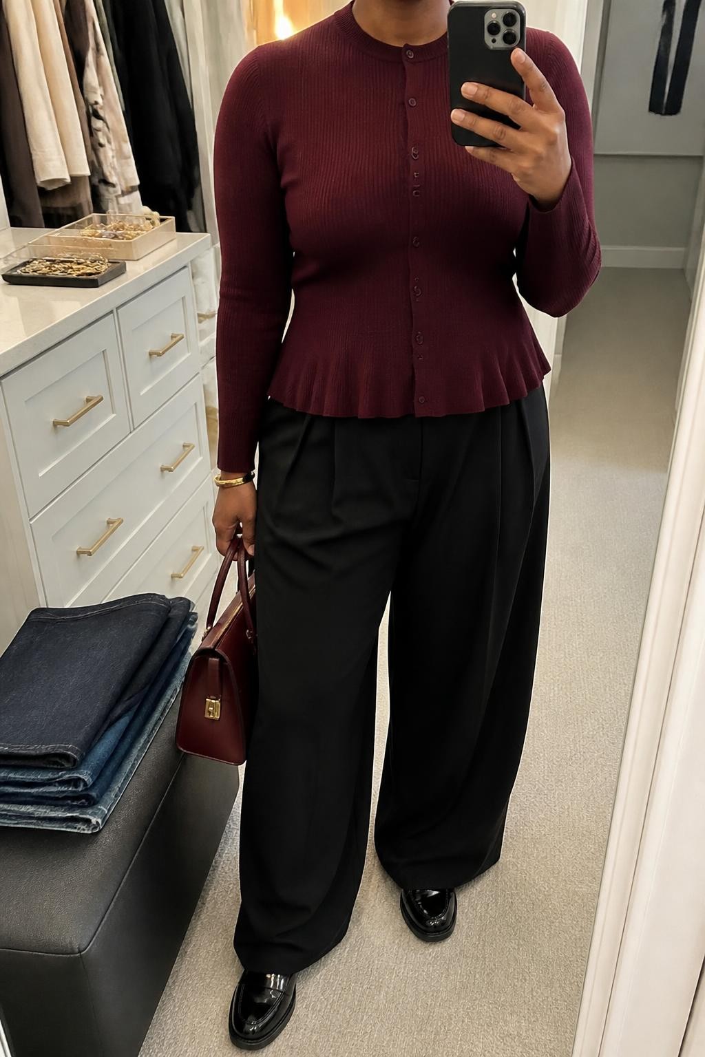 mirror selfie of a Black midsize woman wearing a fully buttoned burgundy fine-knit peplum cardigan, black high-waisted wide-leg trousers, black patent leather loafers, an oxblood structured handbag carried at her side, and a slim gold cuff bracelet, clearly showing the exact clothing items, colors, fabrics, shoes, bag, and bracelet listed in the Outfit Pieces section, holding a phone in front of a mirror, cropped at NECK so the face does not show, relaxed confident selfie pose in a walk-in closet mirror, soft indoor lighting, with folded denim jeans on a bench and a jewelry tray on the vanity, realistic knit and trouser textures, naturally imperfect mirror photo, latest iPhone photo quality
