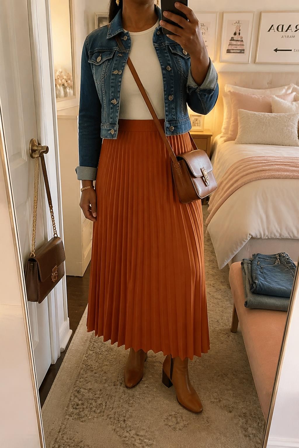 Mirror selfie of a Black woman wearing a rust-orange pleated maxi skirt, cropped medium-wash denim jacket over a fitted ivory crew-neck top, tan leather ankle boots, cognac crossbody bag, and small gold drop earrings, clearly showing the exact clothing items, colors, fabrics, and accessories listed, holding a phone in front of a feminine-themed bedroom mirror, cropped at the neck so the face does not show, relaxed casual selfie pose, evening golden-hour light, handbag hanging on a door handle and folded denim jeans on a bench, casual realistic composition, latest iPhone photo quality.