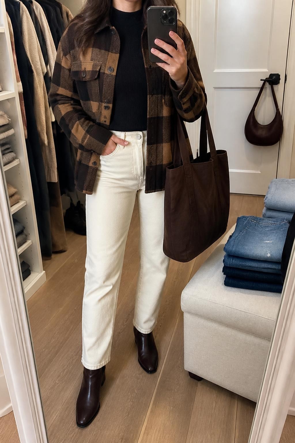 mirror selfie of a woman wearing an open chocolate and camel plaid wool-blend shacket over a black fitted mock-neck knit top, cream high-rise straight-leg jeans, dark brown leather ankle boots, and a chocolate canvas tote bag with no text, clearly showing the exact clothing items, colors, plaid pattern, fabrics, boots, and bag listed in the Outfit Pieces section, holding a phone in front of a mirror, cropped at NECK so the face does not show, relaxed casual selfie pose in a walk-in closet mirror, soft indoor lighting, with folded denim jeans on a bench and a handbag hanging on a door handle, realistic wool, knit, denim, leather, and canvas textures, latest iPhone photo quality