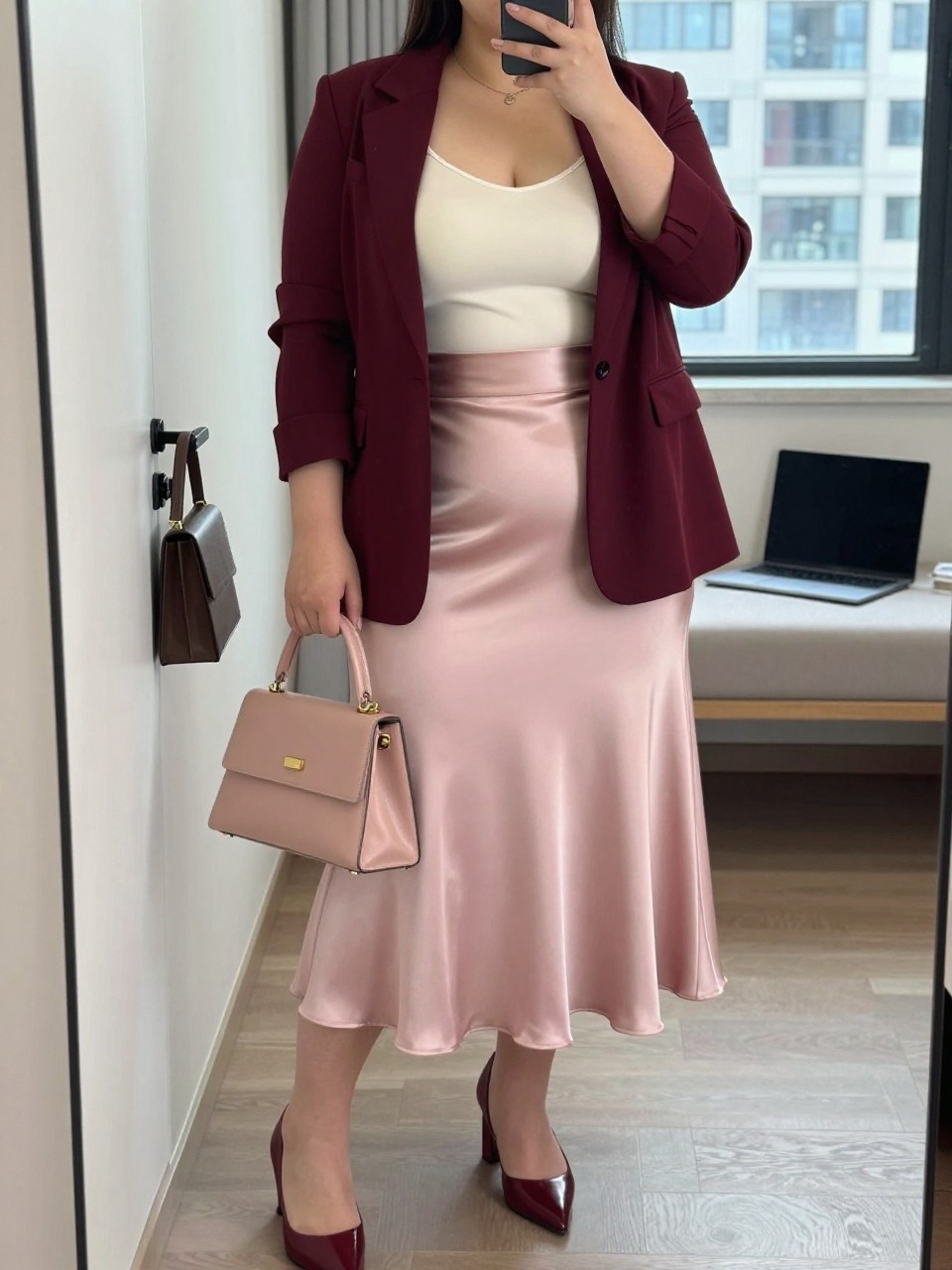 Mirror selfie of a plus size woman wearing a blush pink high-waisted satin midi skirt with a softly flared shape, a fitted ivory scoop-neck camisole with wide straps, a burgundy tailored blazer worn open, burgundy pointed block heels, and a structured blush handbag, clearly showing the exact clothing items, blush satin, ivory top, burgundy blazer, footwear, and accessories listed, holding a phone in front of a mirror, cropped at the neck so her face is hidden, relaxed elegant selfie stance, in a stylish city apartment with natural window light, a handbag hanging on a door handle and a laptop resting on a bench, realistic office celebration outfit, latest iPhone photo quality.