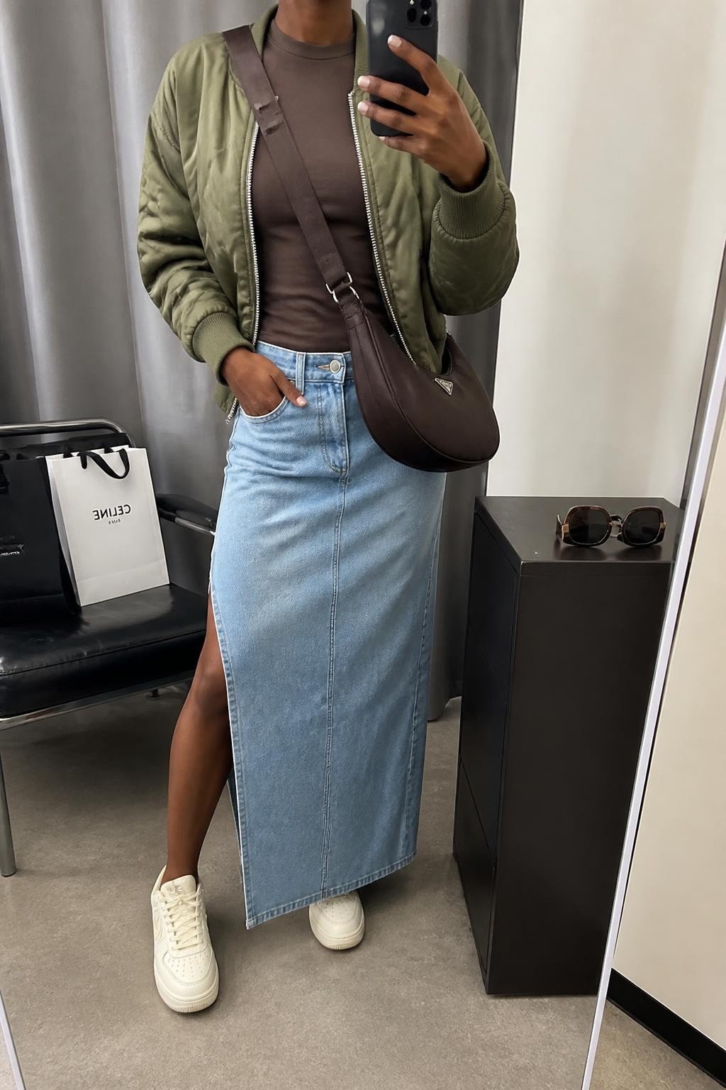 Mirror selfie of a Black woman wearing a light-wash denim maxi skirt with a side slit, fitted chocolate-brown crew-neck top, cropped olive-green quilted bomber jacket, cream leather sneakers, and a dark brown nylon crossbody bag, clearly showing the exact clothing items, colors, fabrics, and accessories listed, holding a phone in front of a fashion studio mirror corner, cropped at the neck so the face does not show, relaxed casual selfie pose, soft indoor lighting, shopping bags beside a chair and sunglasses placed on a dresser, realistic casual scene, latest iPhone photo quality.