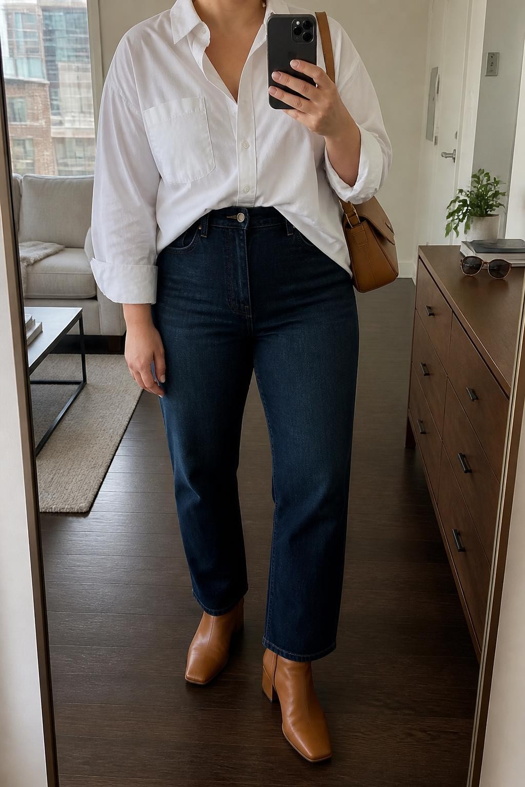 Mirror selfie of a plus size woman wearing an oversized white cotton button-down shirt with the front section tucked into dark indigo high-waisted straight-leg jeans, tan leather ankle boots with low block heels, a structured camel shoulder bag, and small gold hoop earrings, clearly showing the exact clothing items, white cotton, dark denim, tan footwear, and accessories listed, holding a phone in front of a mirror, cropped at the neck so the face does not show, relaxed casual selfie pose, in a stylish city apartment with natural window light, sunglasses placed on a dresser and a small plant nearby, natural unstaged appearance, latest iPhone photo quality.